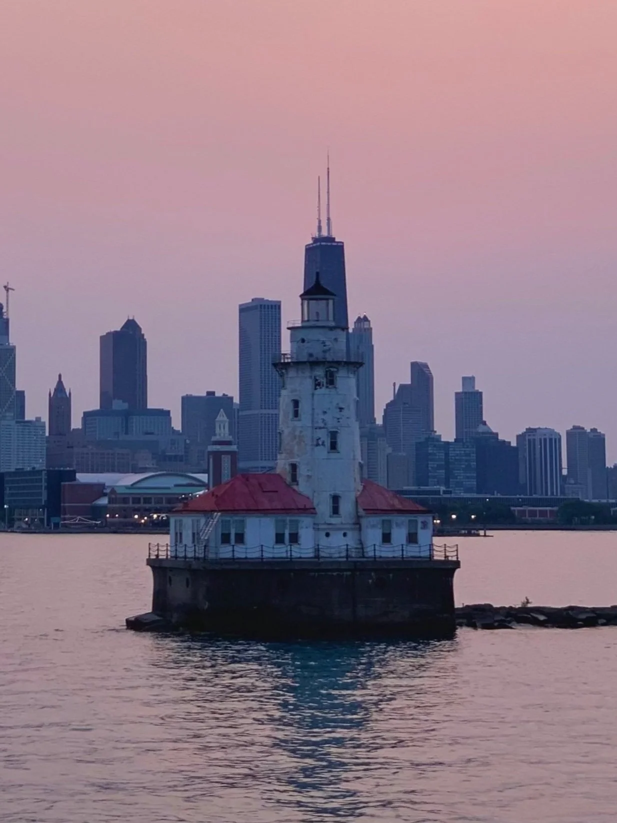 A lighthouse on a small island in the water with Chicago skyline in the background during sunset.