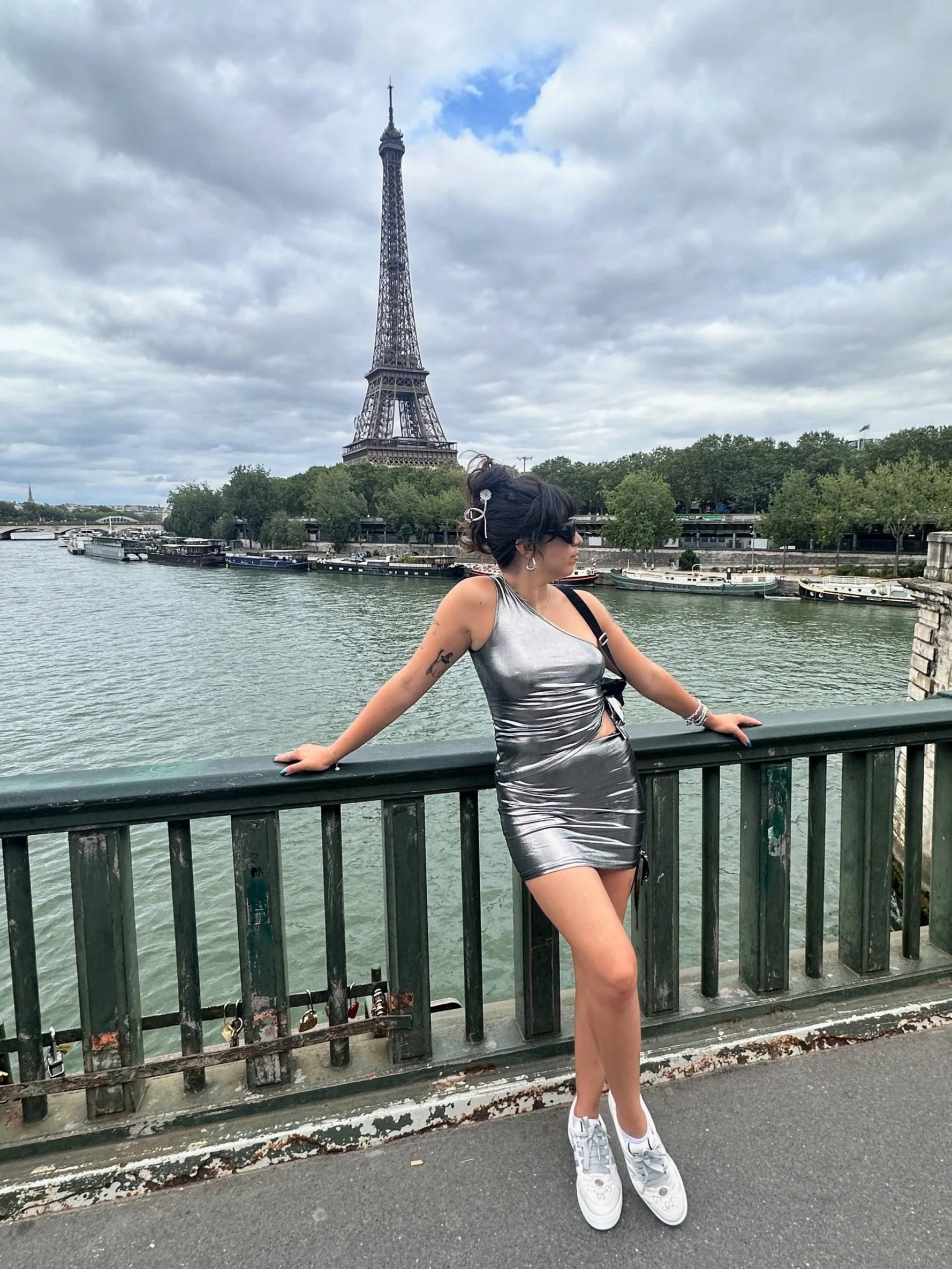 A woman dressed in a metallic silver dress and white sneakers leaning on a bridge railing along the Seine River with the Eiffel Tower in the background on a cloudy day in Paris.