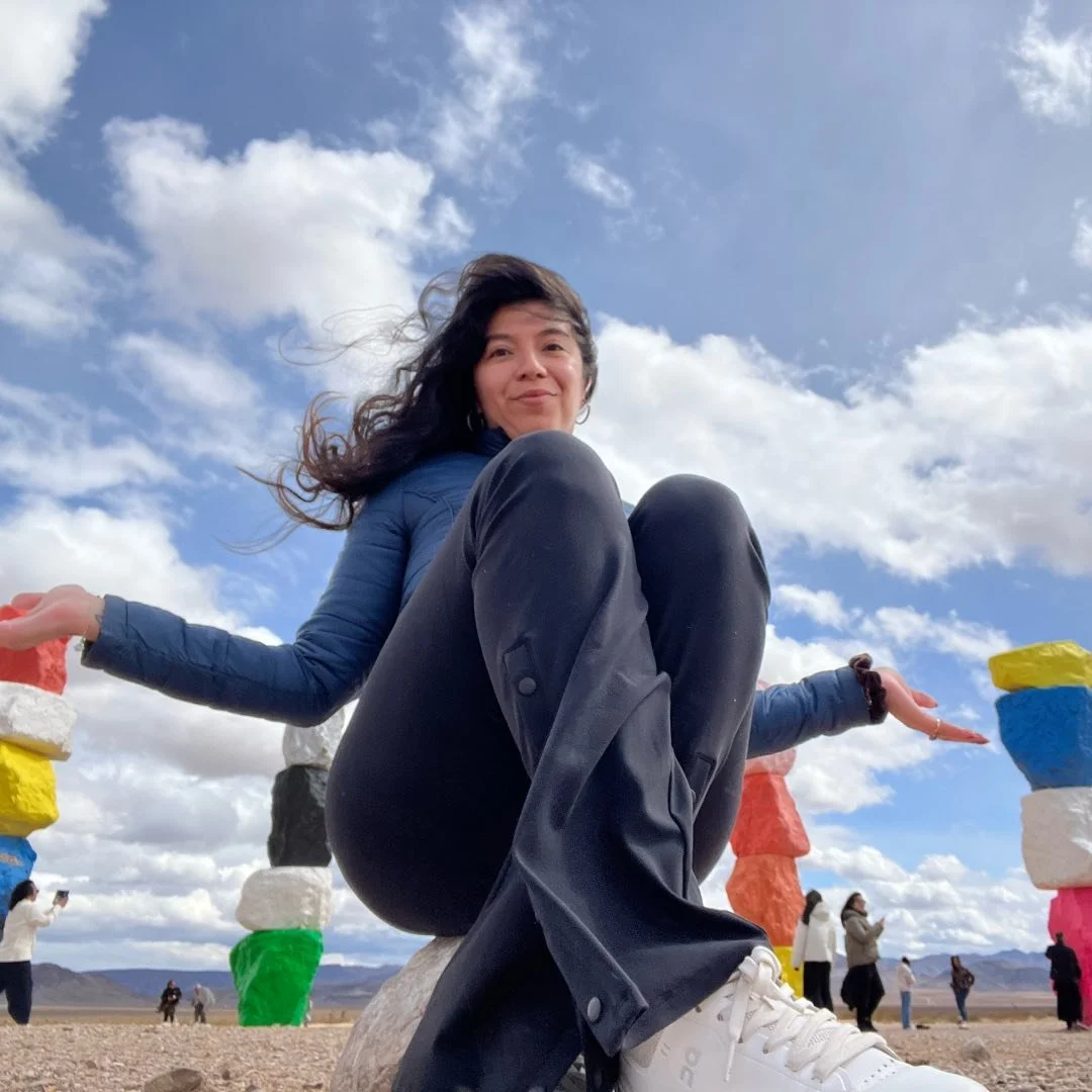 A woman sitting on a stone in an outdoor desert landscape with colorful rock formations in the background, under a partly cloudy sky.
