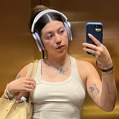 Woman taking a selfie in an elevator with headphones on, wearing a white tank top, carrying a beige bag, and displaying tattoos on her arm and wrist.