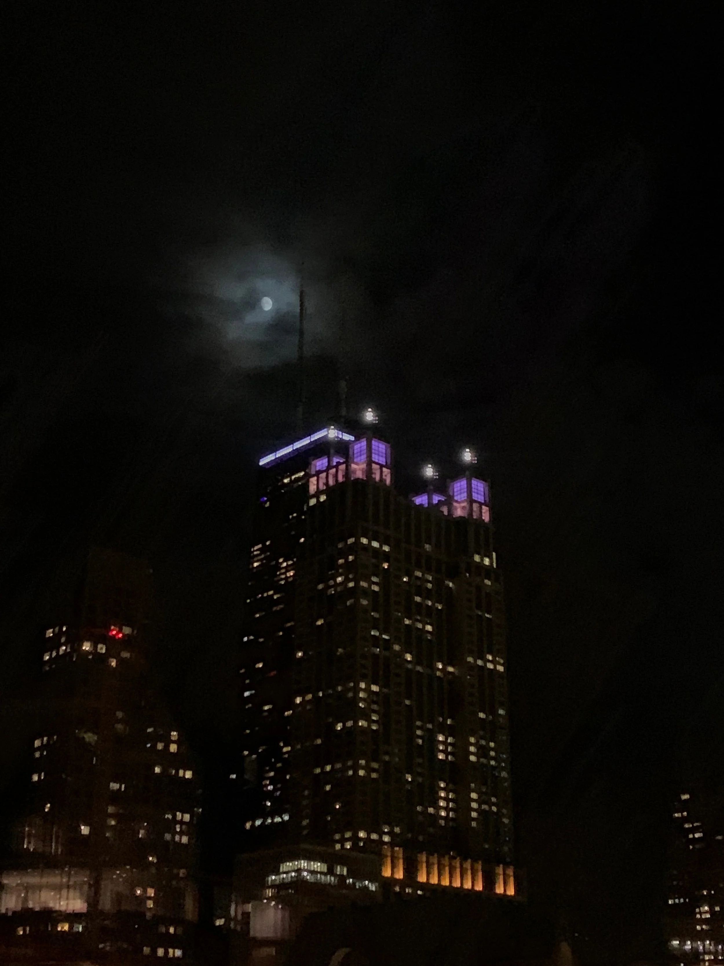 Nighttime photograph of a tall building with purple and pink lights on the top, with the moon visible behind clouds in a dark sky.