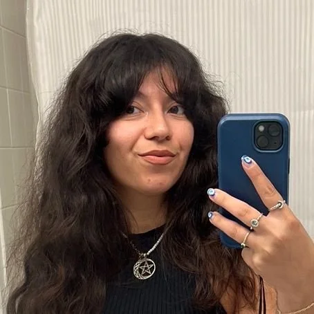 Self-portrait of a woman with long, wavy dark hair taking a mirror selfie with a blue phone in a room with cream-colored striped wallpaper.