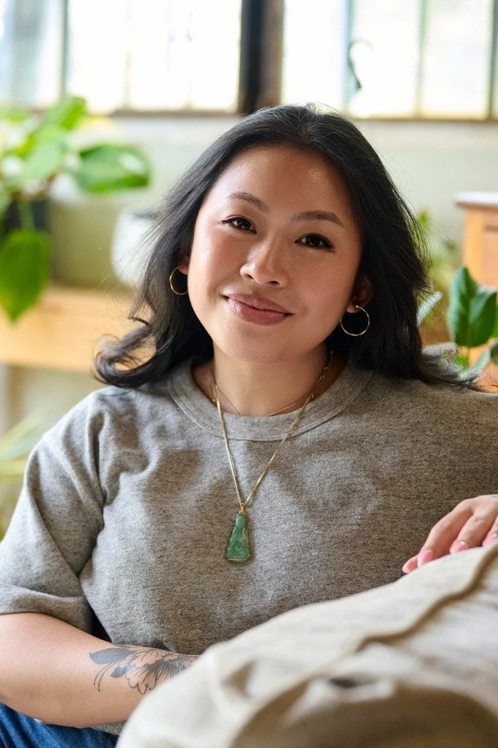 Marya with dark hair, wearing a gray shirt and jewelry, sitting indoors with potted plants and a window in the background.