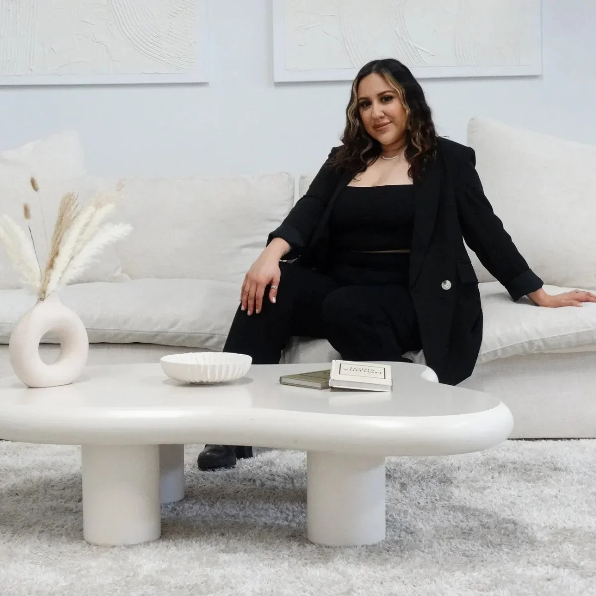 Woman with dark hair and a black outfit sitting on a white sofa in a modern, minimalist living room with neutral decor.