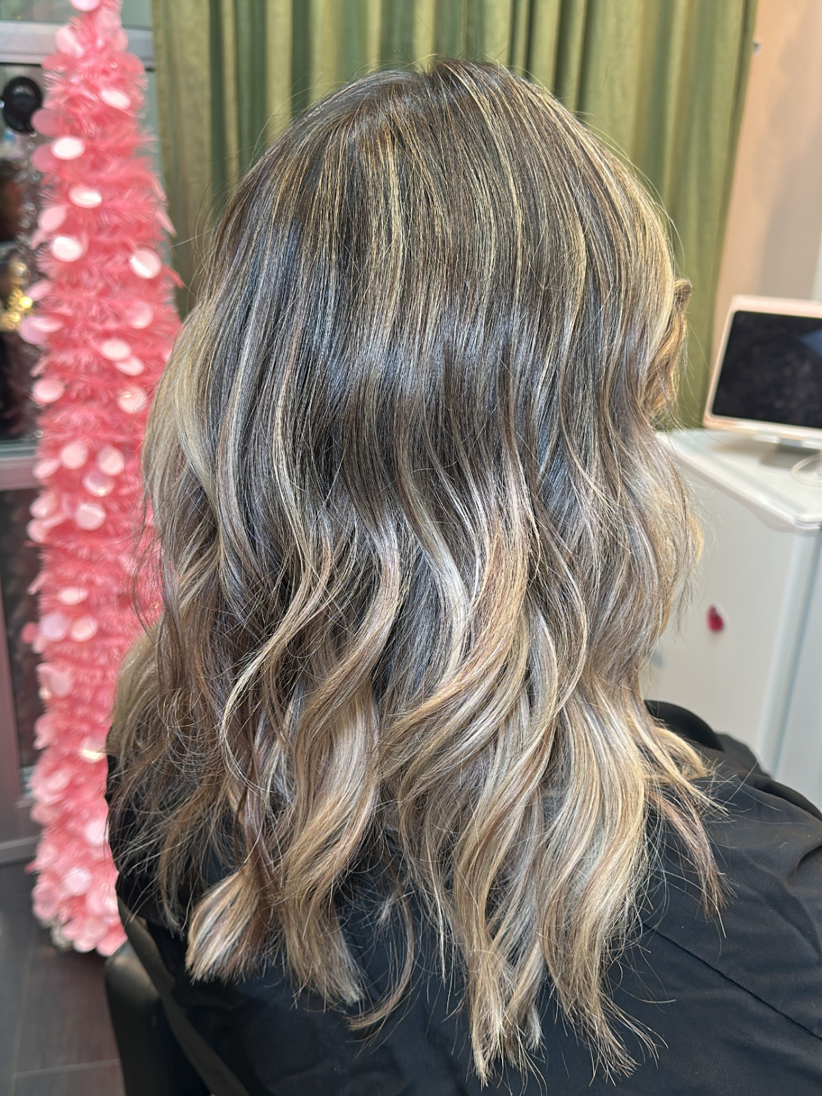 Back of a clients head with shoulder-length wavy blonde and brown hair. In the background, there is a pink feathered decoration and some green curtains.