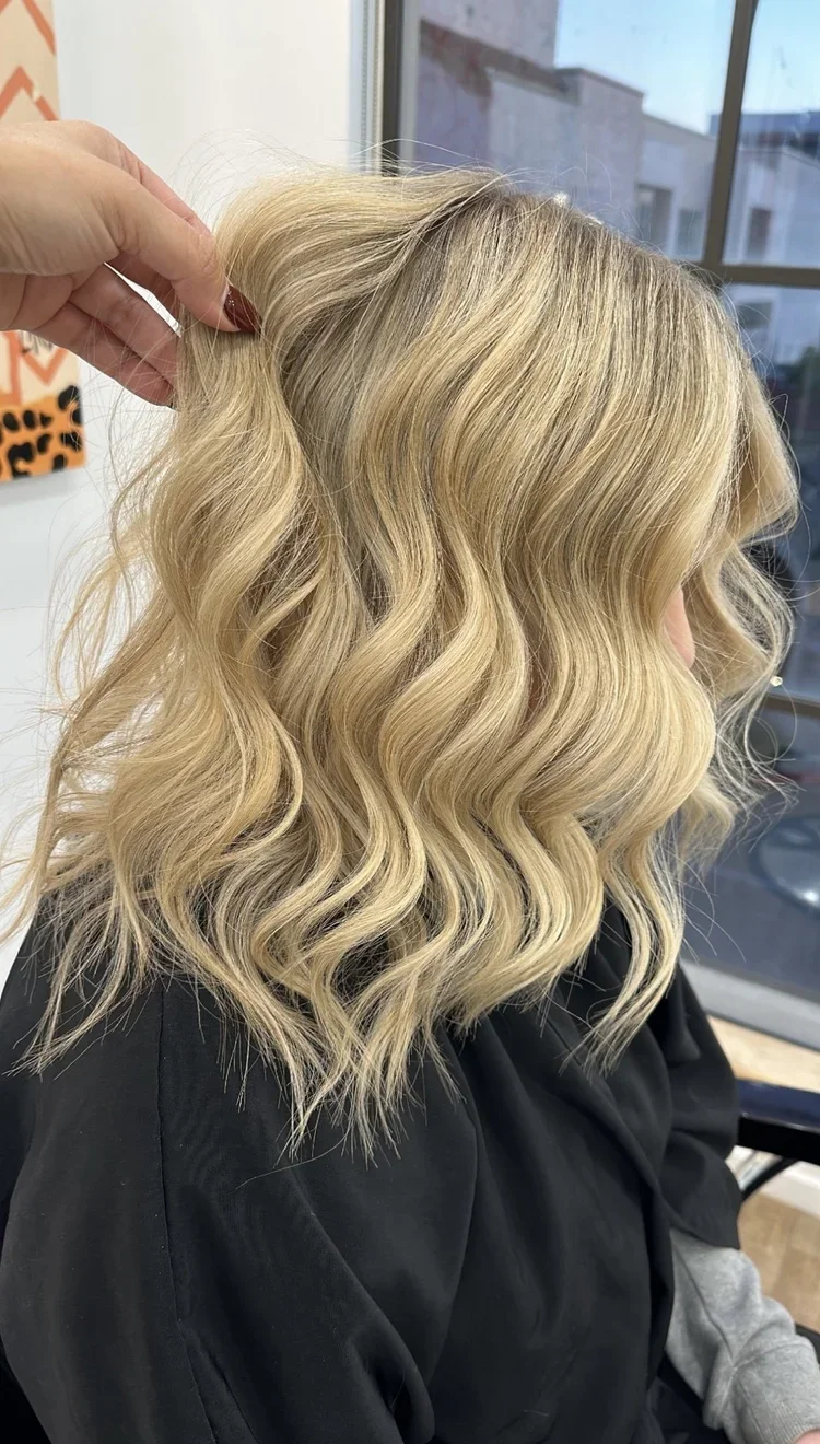 Close-up of a woman's blonde, wavy hair being styled in a salon with a window in the background.