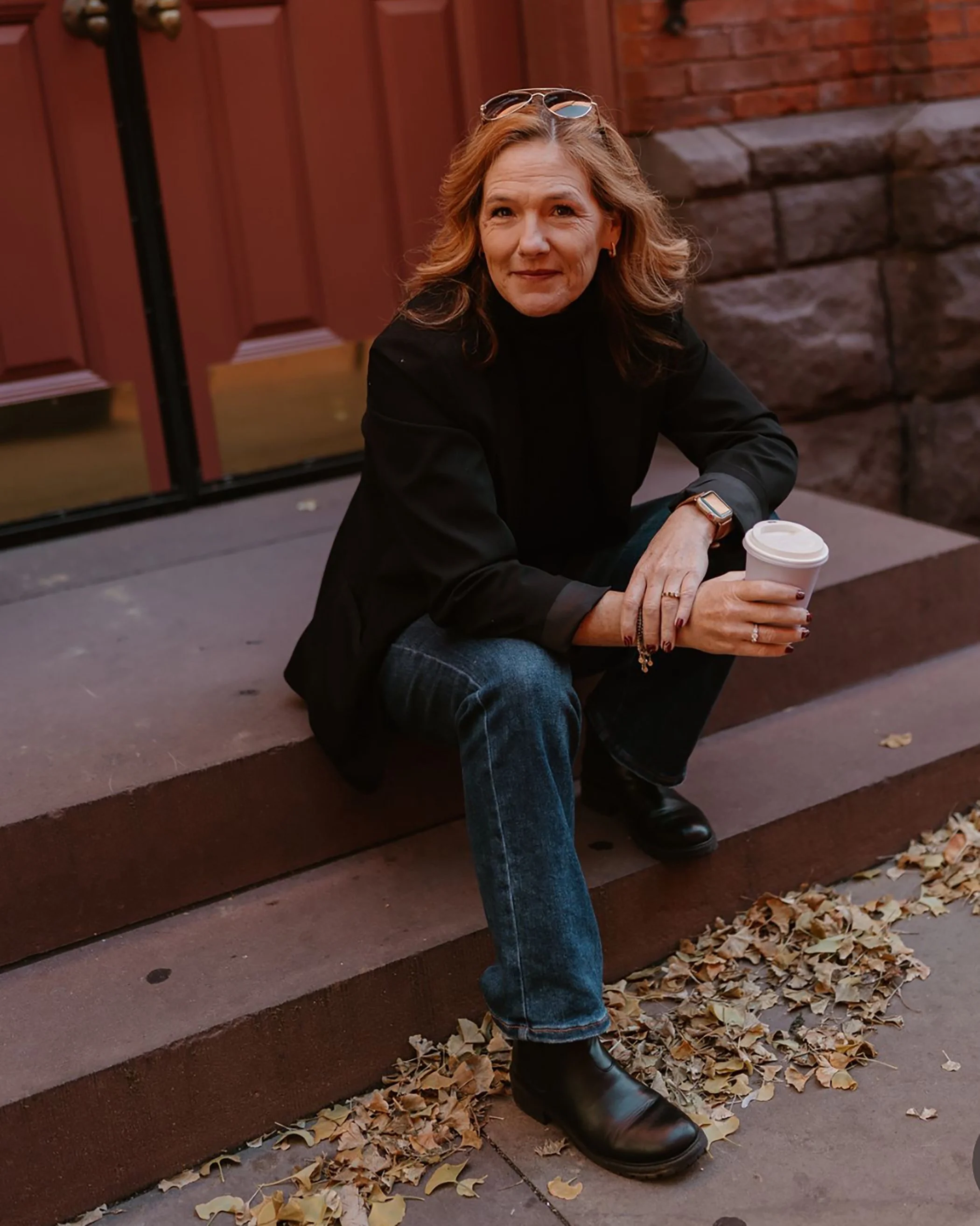 Heather Pfeifer sitting on a step outside, holding a coffee cup, wearing  a black jacket, jeans, black boots, and glasses on her head, with autumn leaves on the ground.