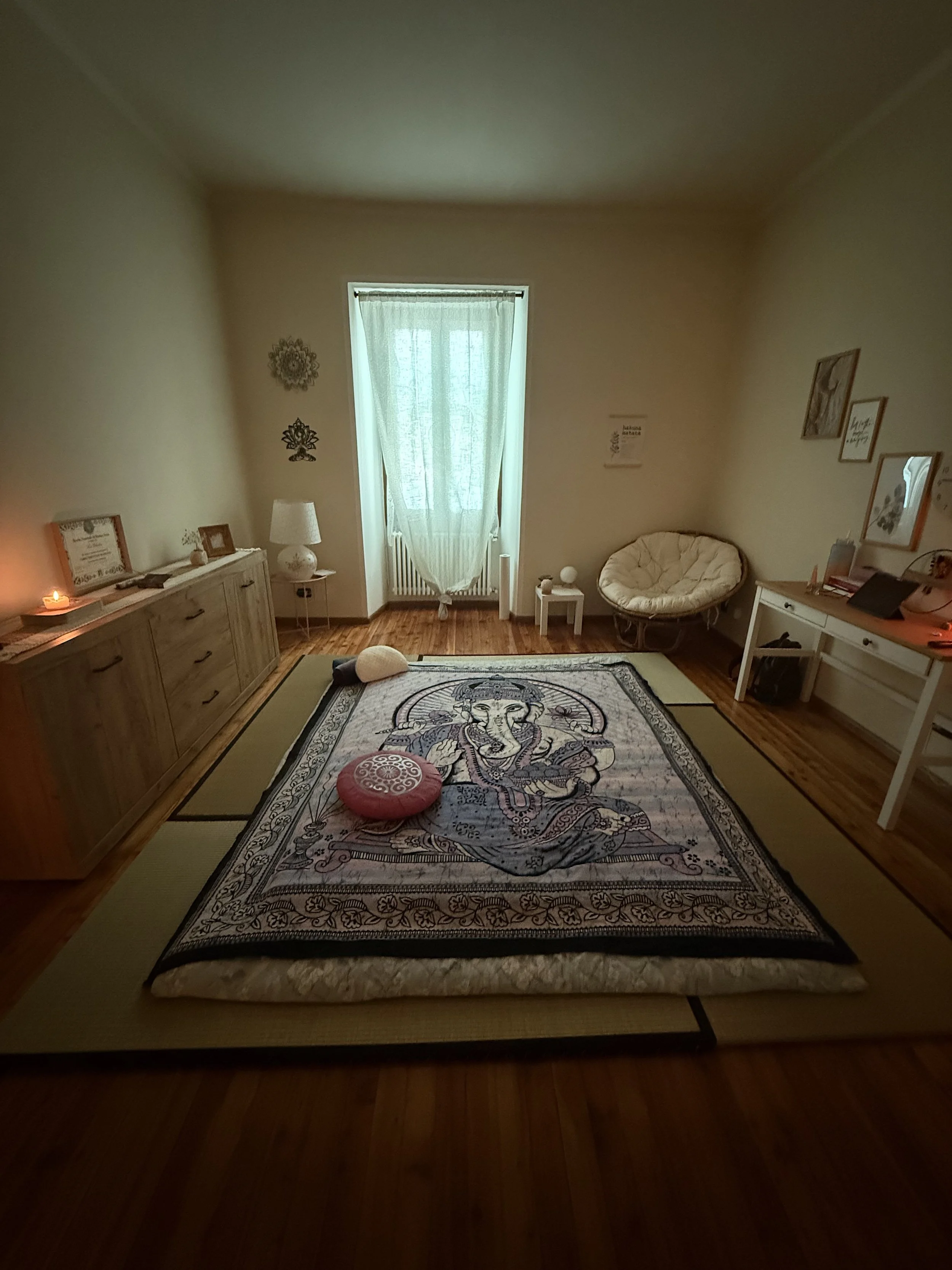 Stanza con tappeto con immagine di Ganesha, cuscini rosa e bianco, mobile con decorazioni, poltrona, scrivania con oggetti e quadro alle pareti, cortile con finestra con tende.