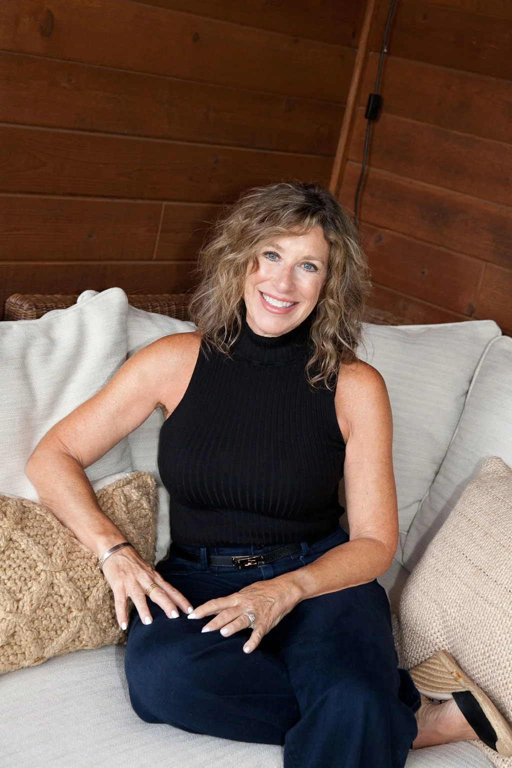 A smiling woman with curly blond hair, wearing a sleeveless black turtleneck top and black pants, sitting on a beige sofa with pillows in a wooden-paneled room.