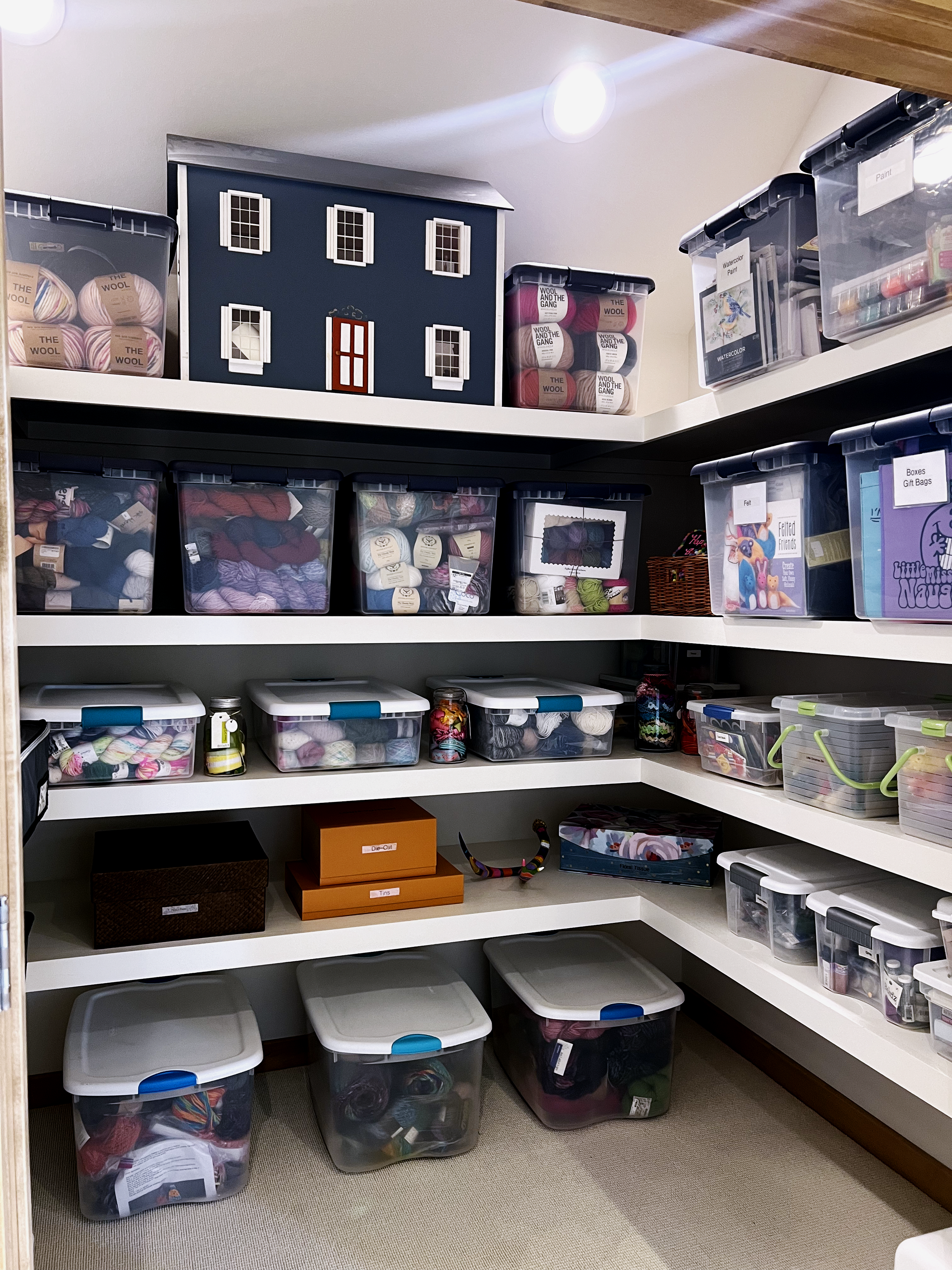 Closet with several shelves filled with transparent plastic storage bins containing yarn and knitting supplies, a decorative dollhouse on the top shelf, and a few small boxes and accessories.