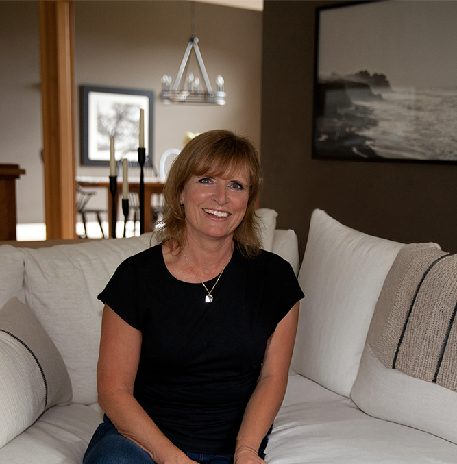 A middle-aged woman with light brown hair is sitting on a beige sectional sofa in a living room, smiling at the camera. She is wearing a black top and a necklace.