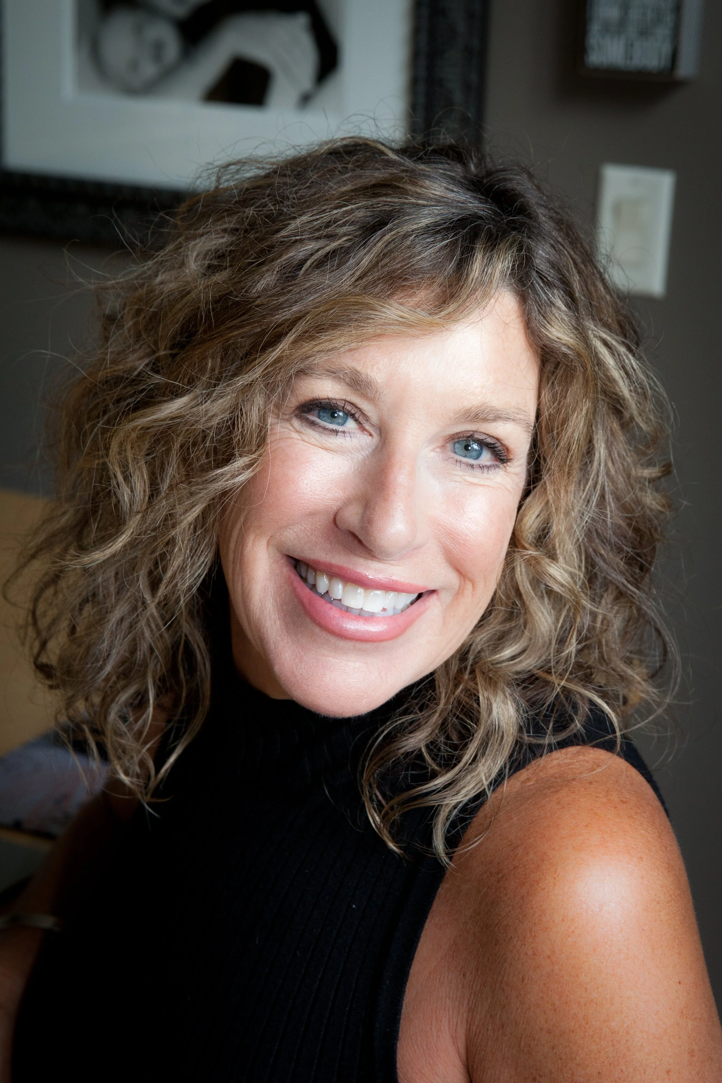 Close-up of a woman with curly, shoulder-length light brown hair, blue eyes, and a bright smile, wearing a black sleeveless top.