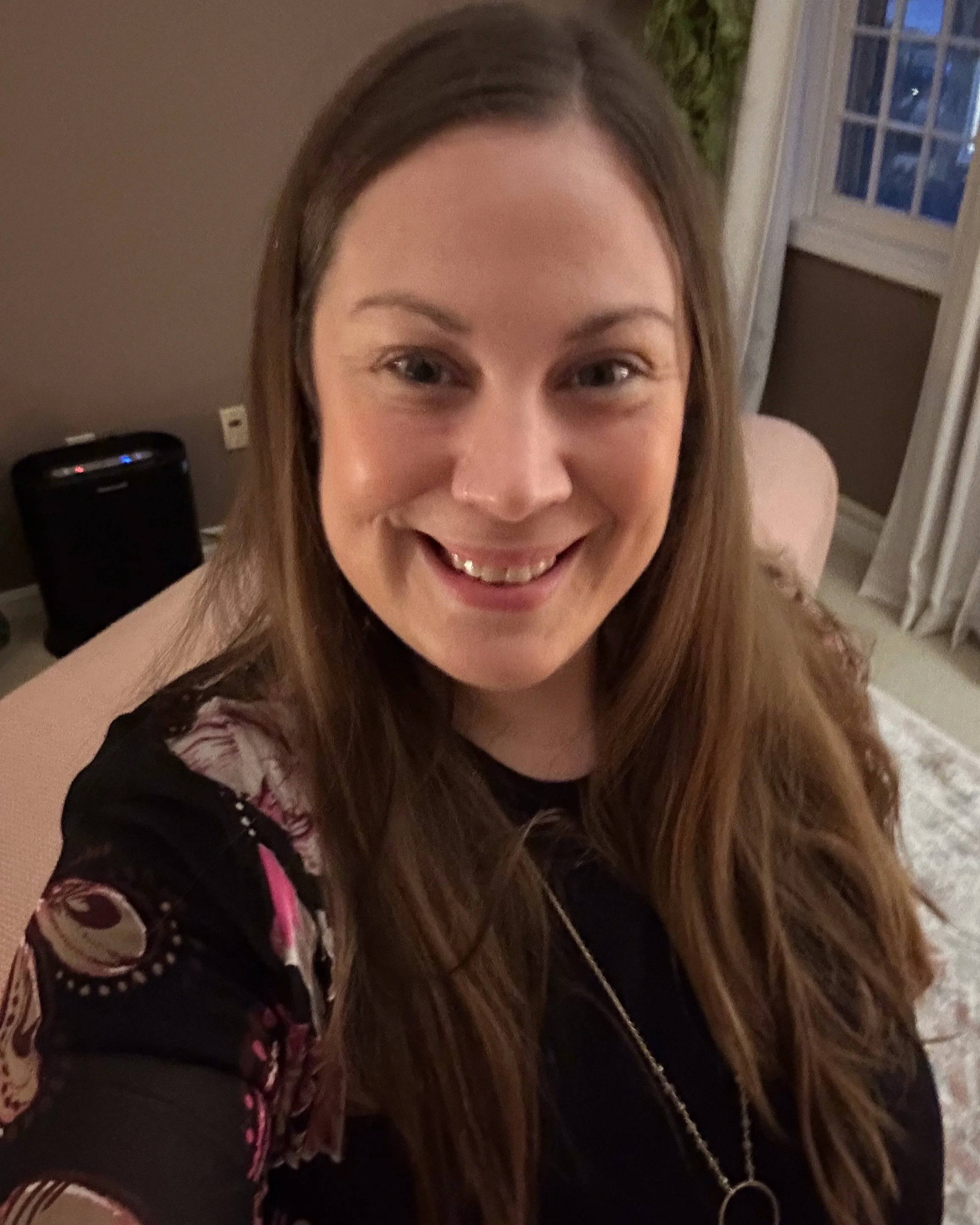 A woman with long brown hair smiling at the camera, sitting in a warmly lit room with a large window and curtains in the background.