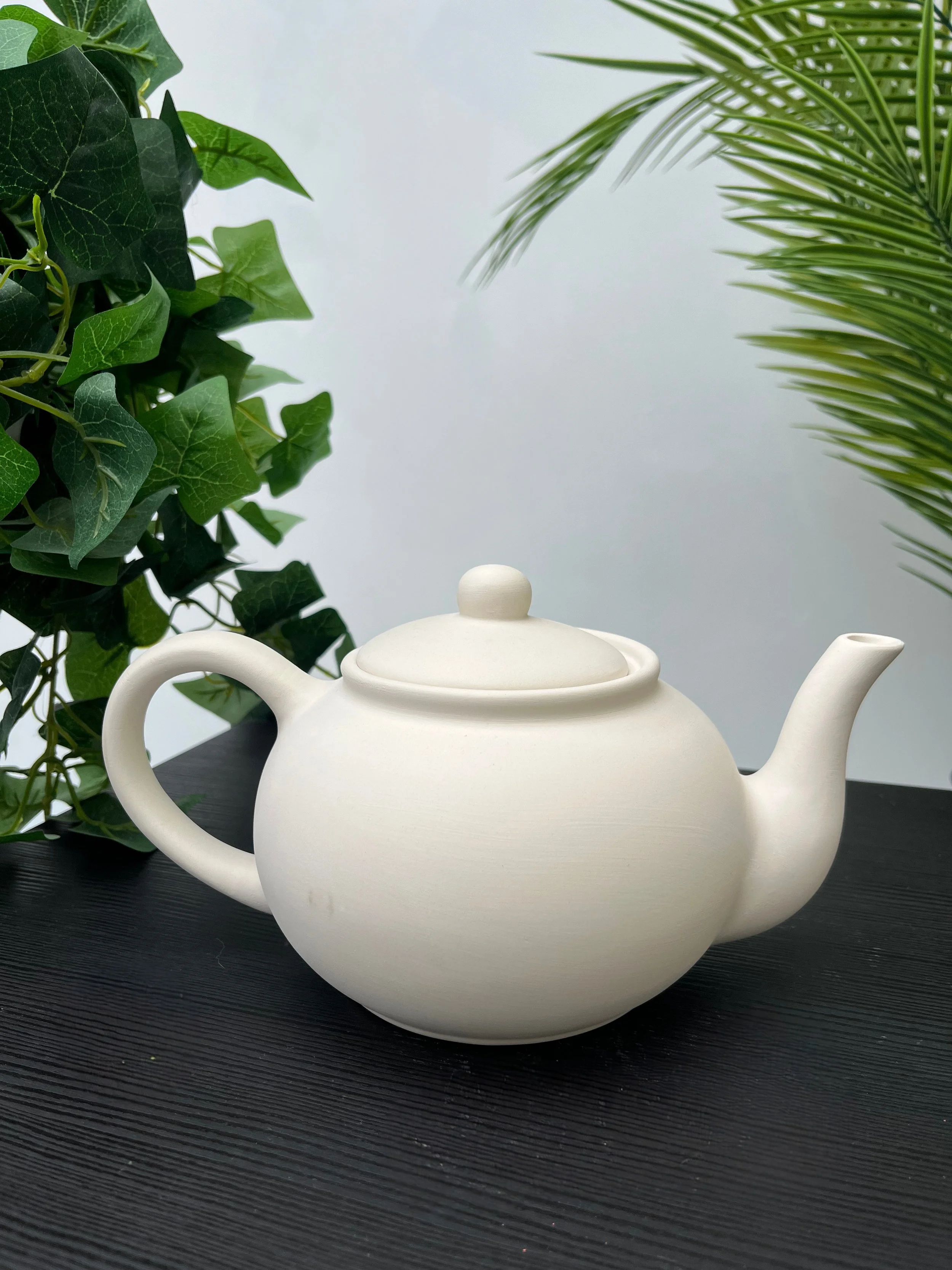 White ceramic teapot on a dark wooden table with green houseplants in the background.