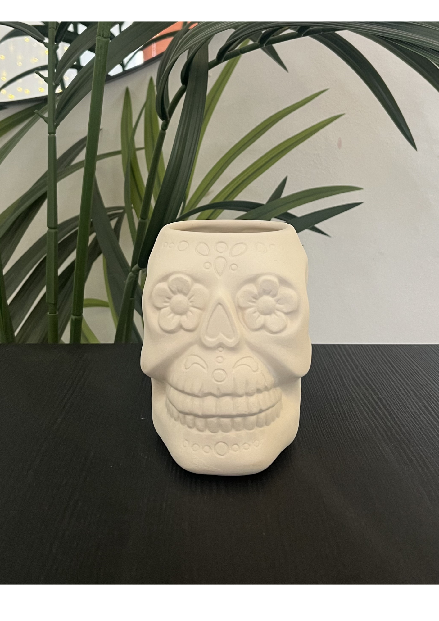 A ceramic mug shaped like a skull with a handle, placed on a dark textured table with green leafy plants in the background.