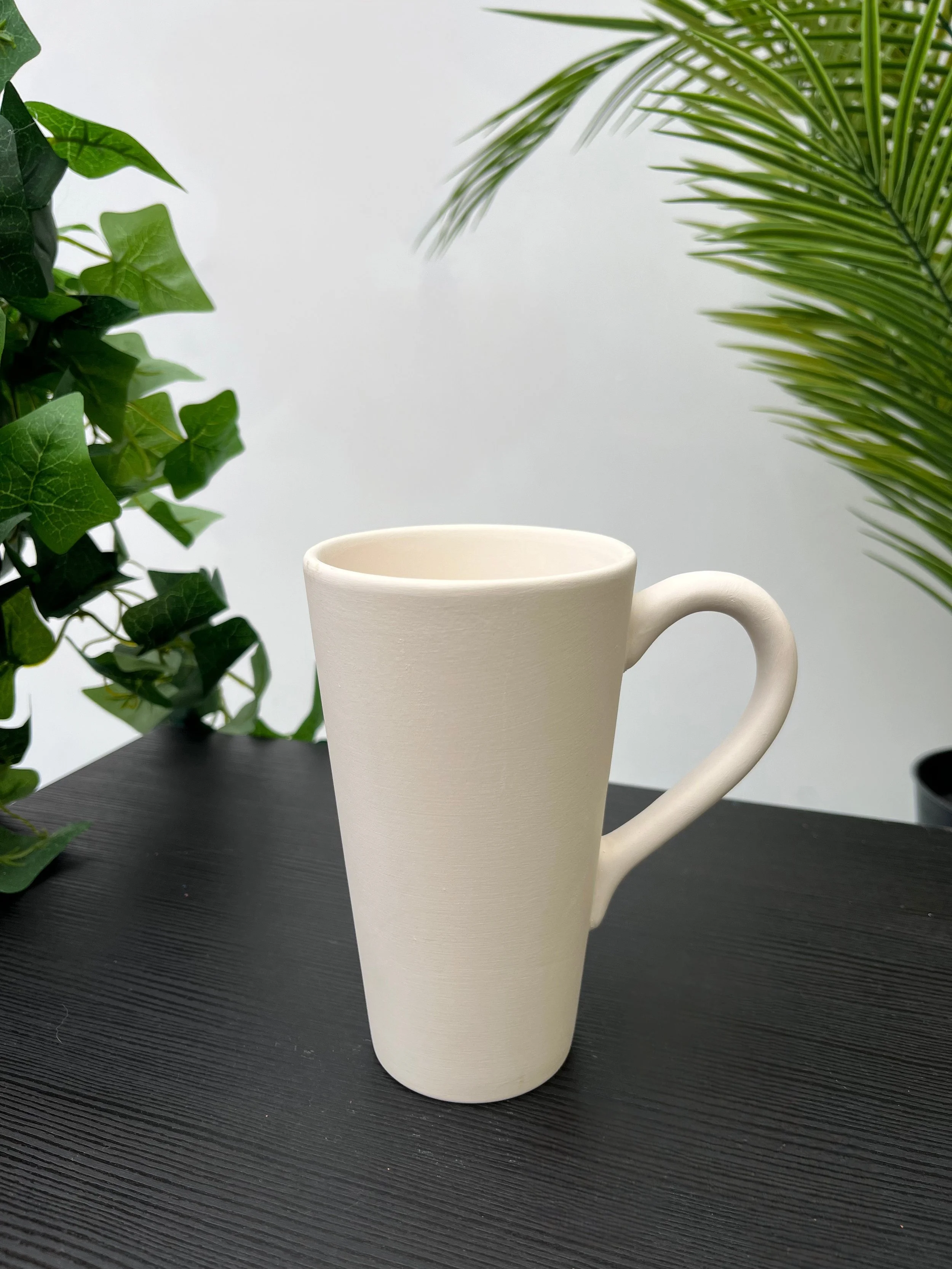 A coffee mug with a unique shape, featuring a rounded handle and an elongated body, placed on a dark wooden table, with a green leafy plant in the background.