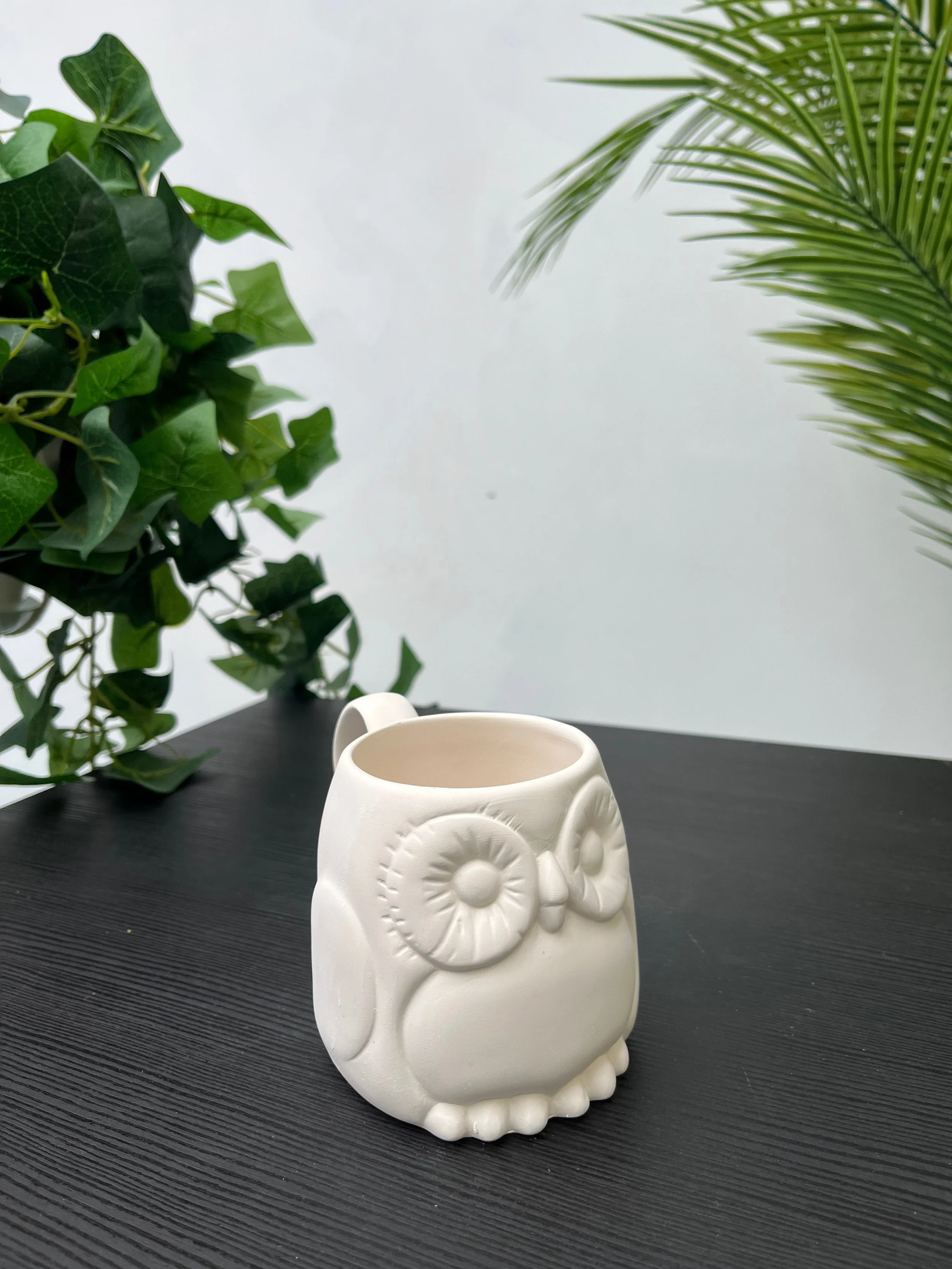 A white owl-shaped ceramic mug on a black surface, with large green leafy plants in the background.