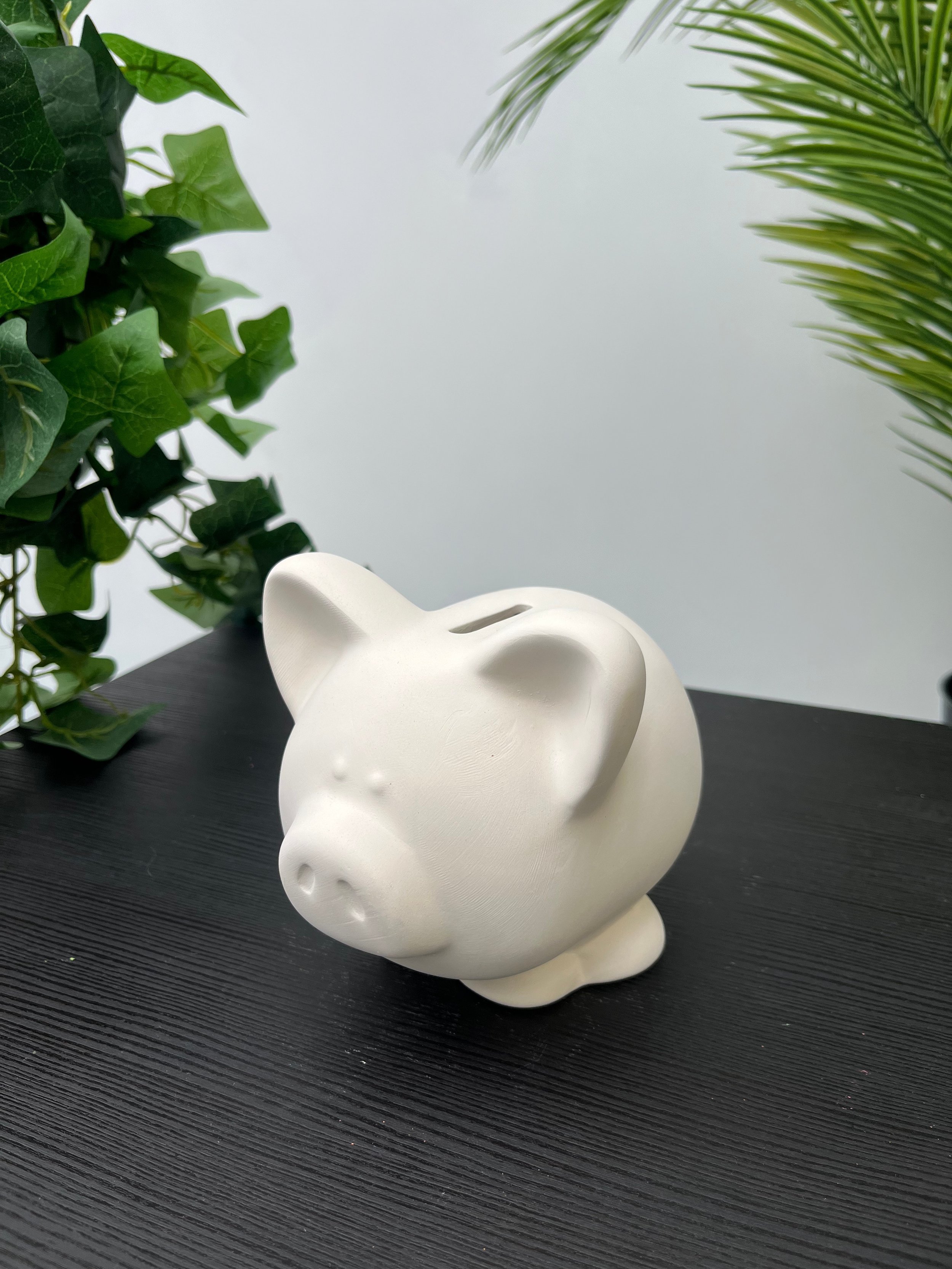 White ceramic piggy bank placed on a dark textured table with green plant leaves in the background.