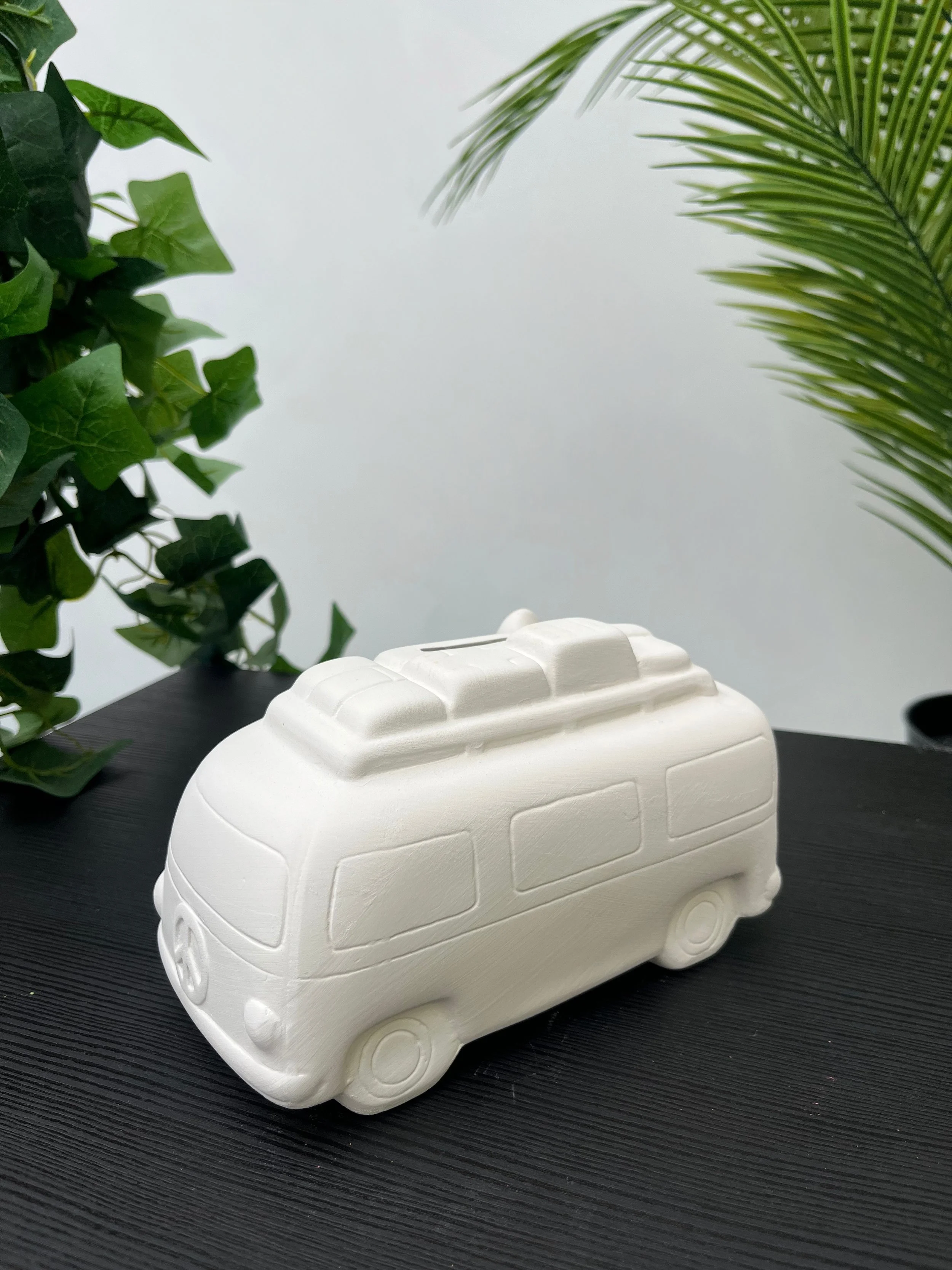 A white ceramic of a VW camper van on a dark wooden surface with green leafy plants in the background.