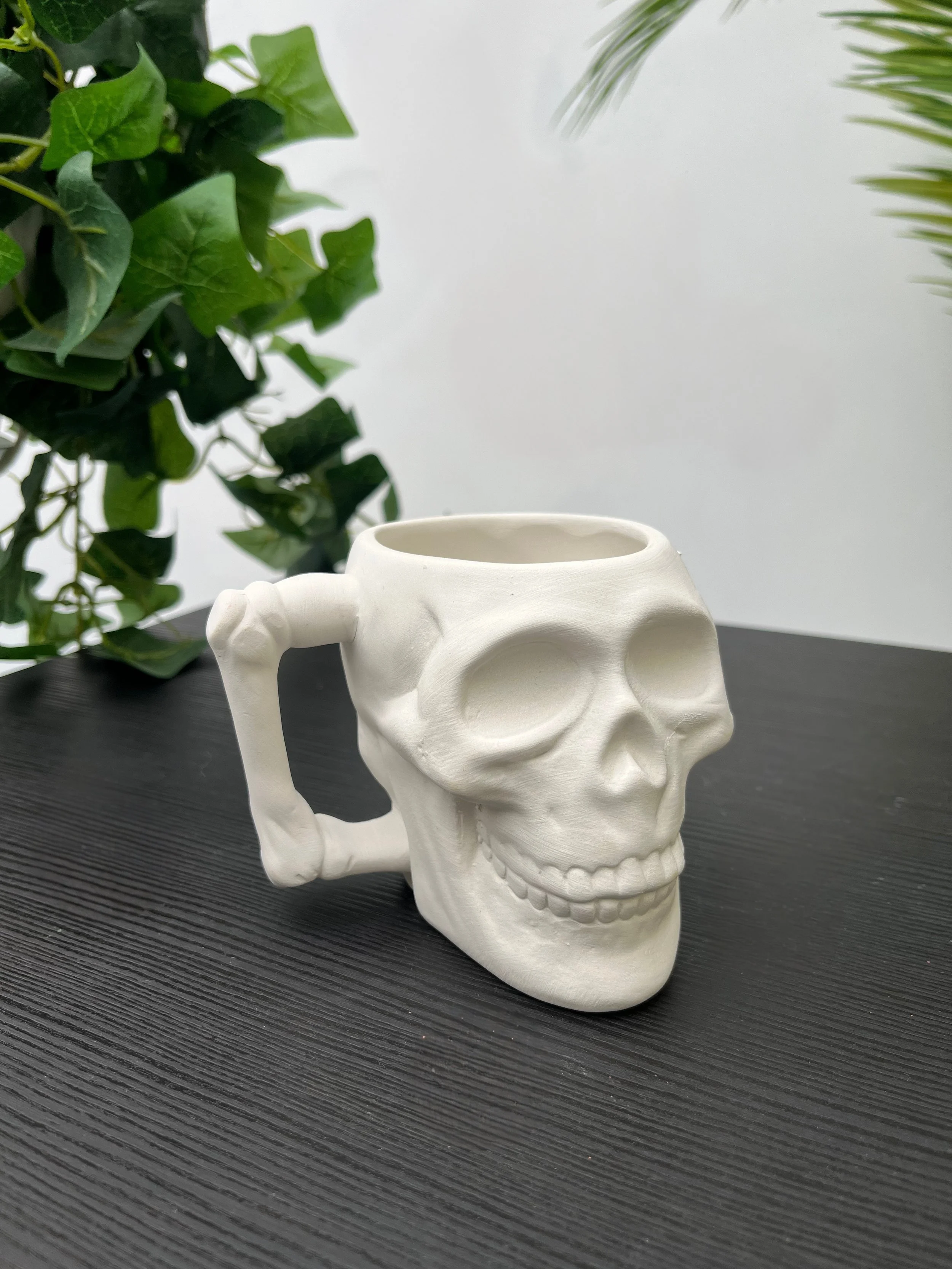 A ceramic mug shaped like a skull with a handle, placed on a dark textured table with green leafy plants in the background.