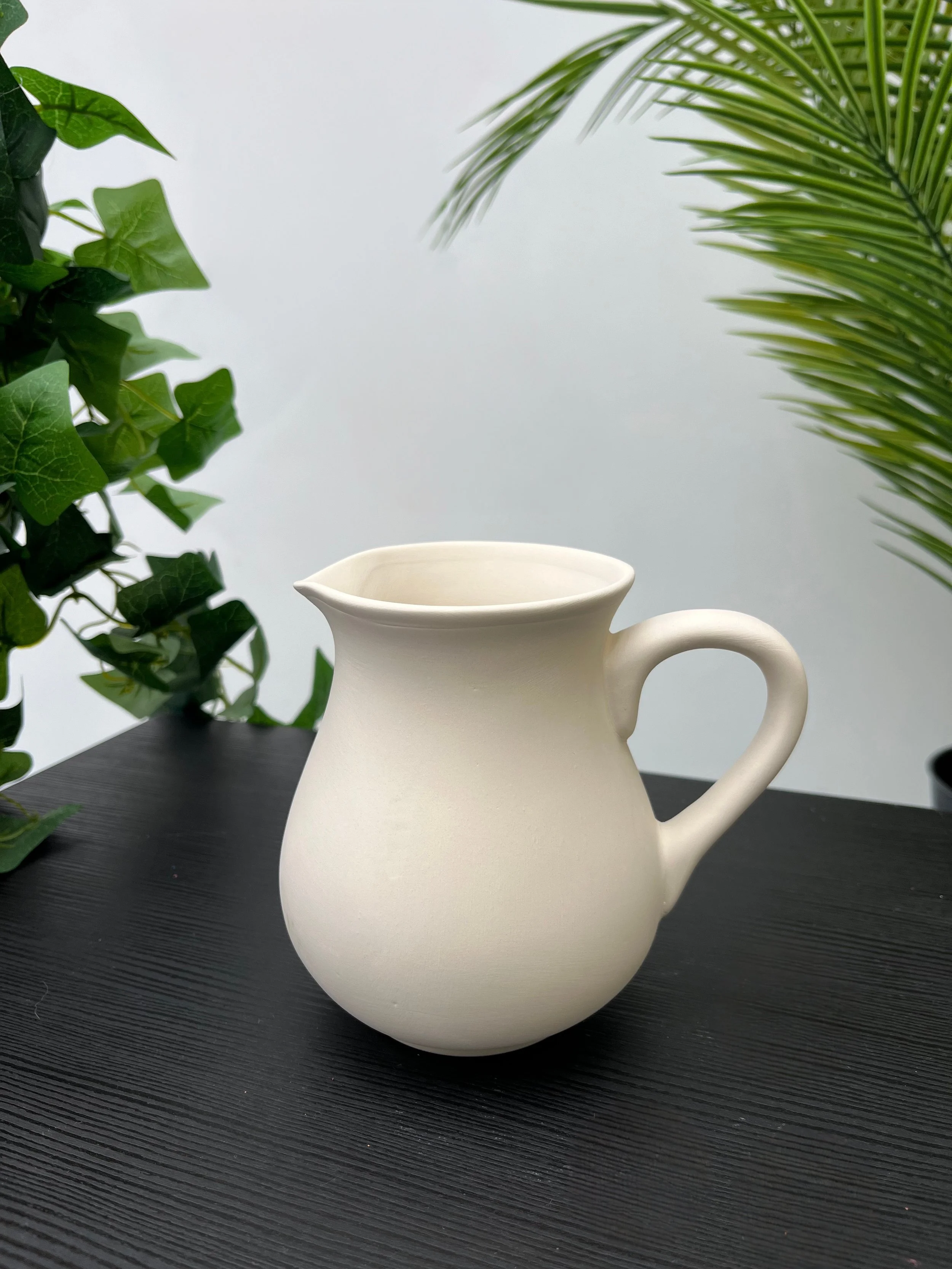 White ceramic pitcher with a handle placed on a black table, with green foliage in the background.