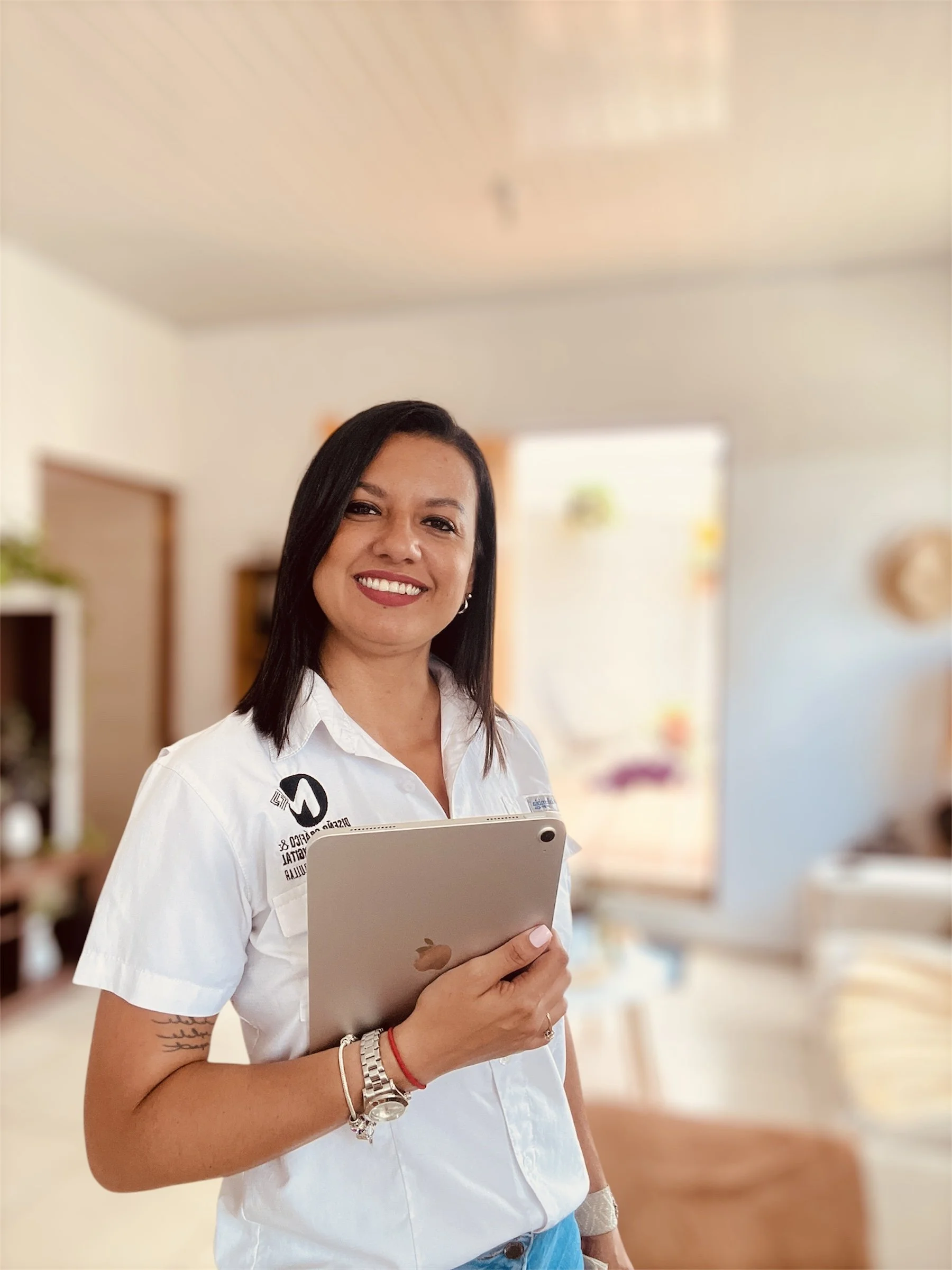 Mujer sonriendo, con cabello negro y camisa blanca, sosteniendo una tableta en un entorno interior con fondo difuso.