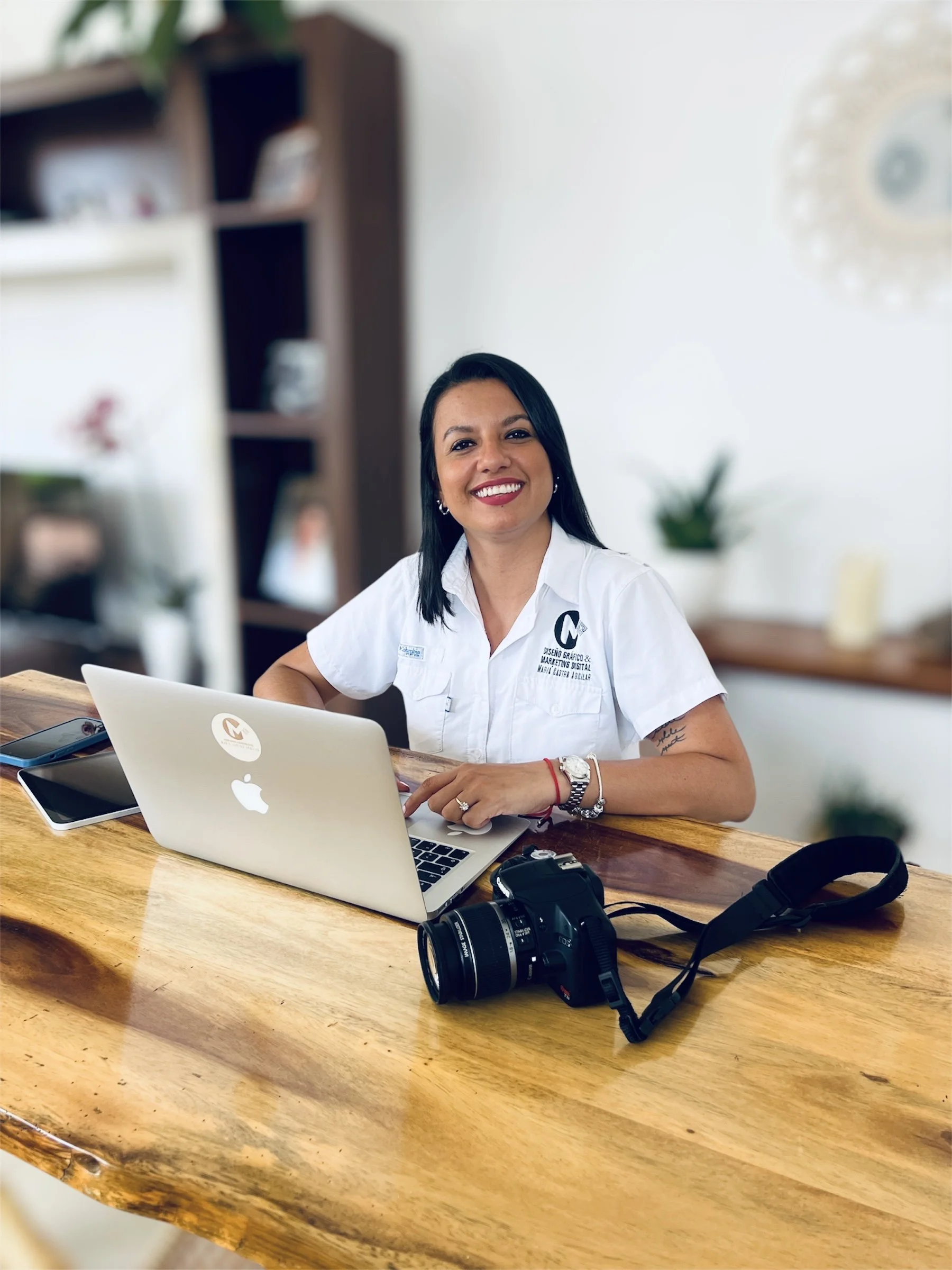 Mujer sonriendo trabajando en un escritorio con una computadora portátil, una cámara y un teléfono móvil.