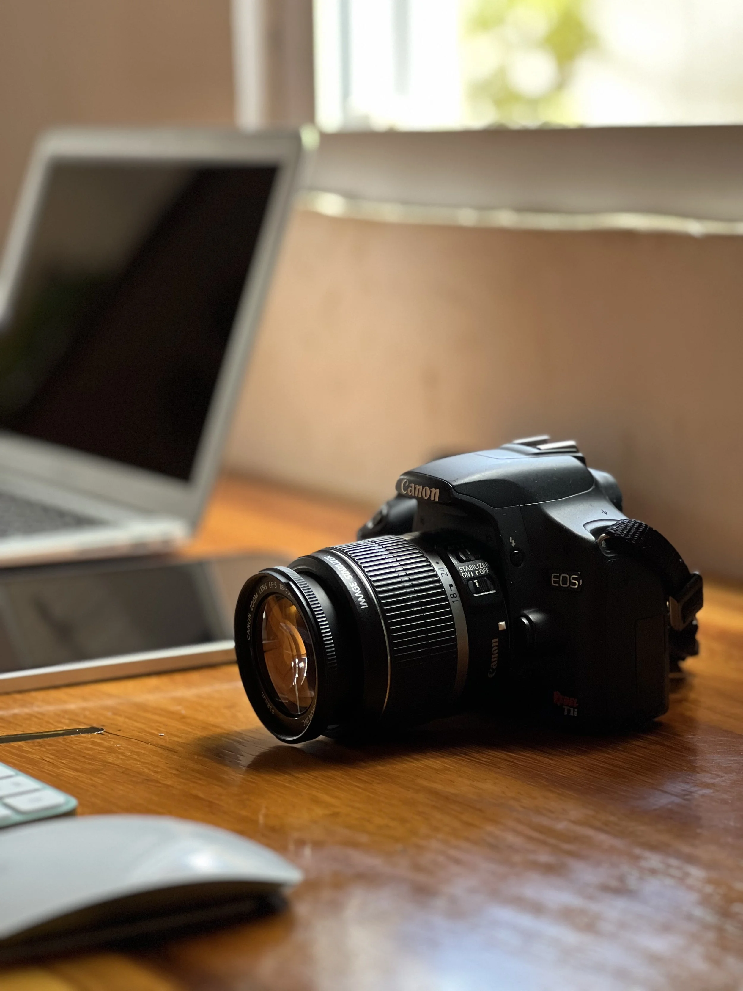 Cámara Canon en un escritorio de madera acompañada de una laptop, mouse y teclado.