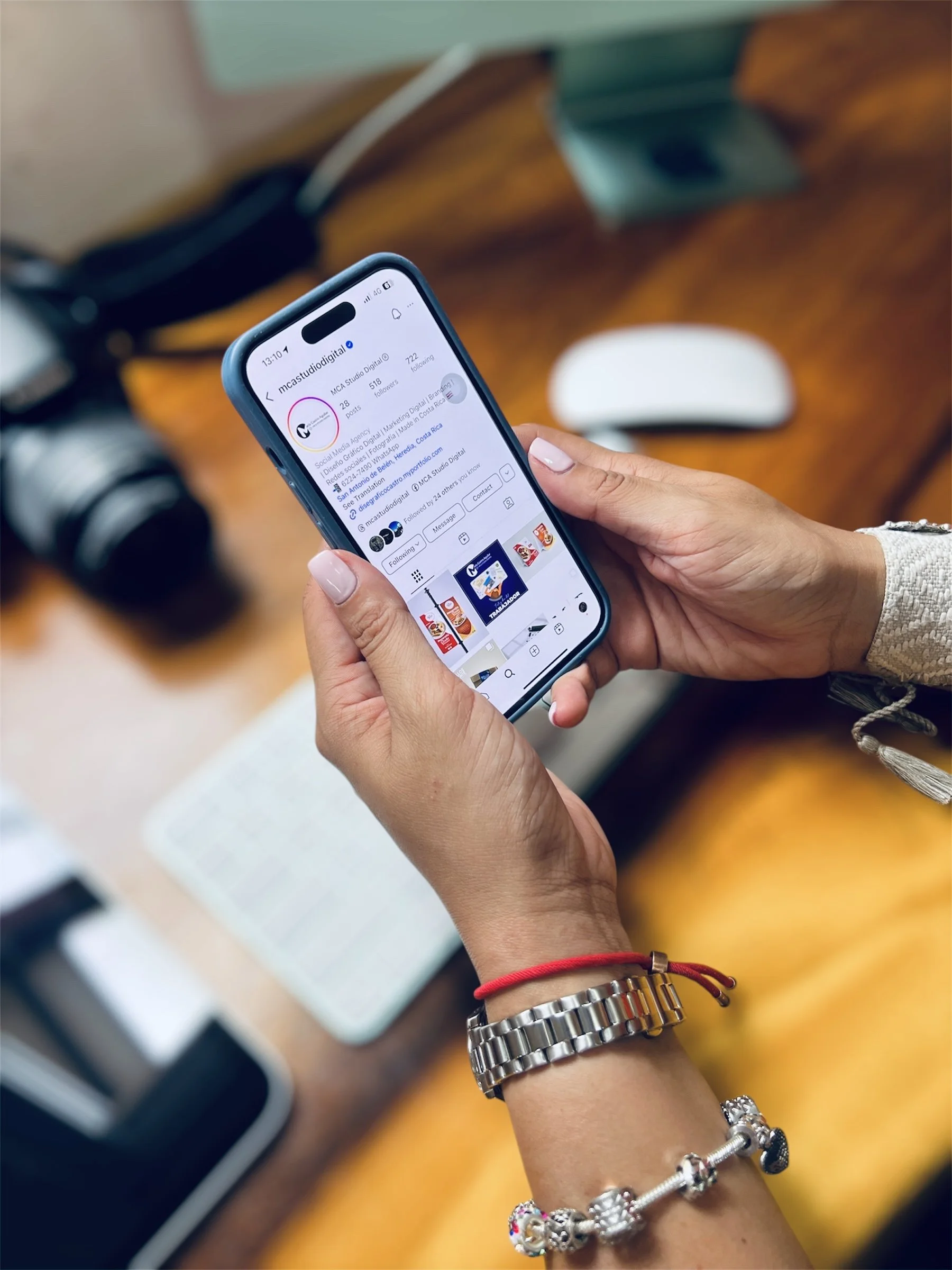 Manos sosteniendo un teléfono móvil que muestra la cuenta de Instagram de @mcastudiodigital en una mesa de madera. La mano lleva una pulsera de reloj de metal, una pulsera de charms y una banda de tela roja.