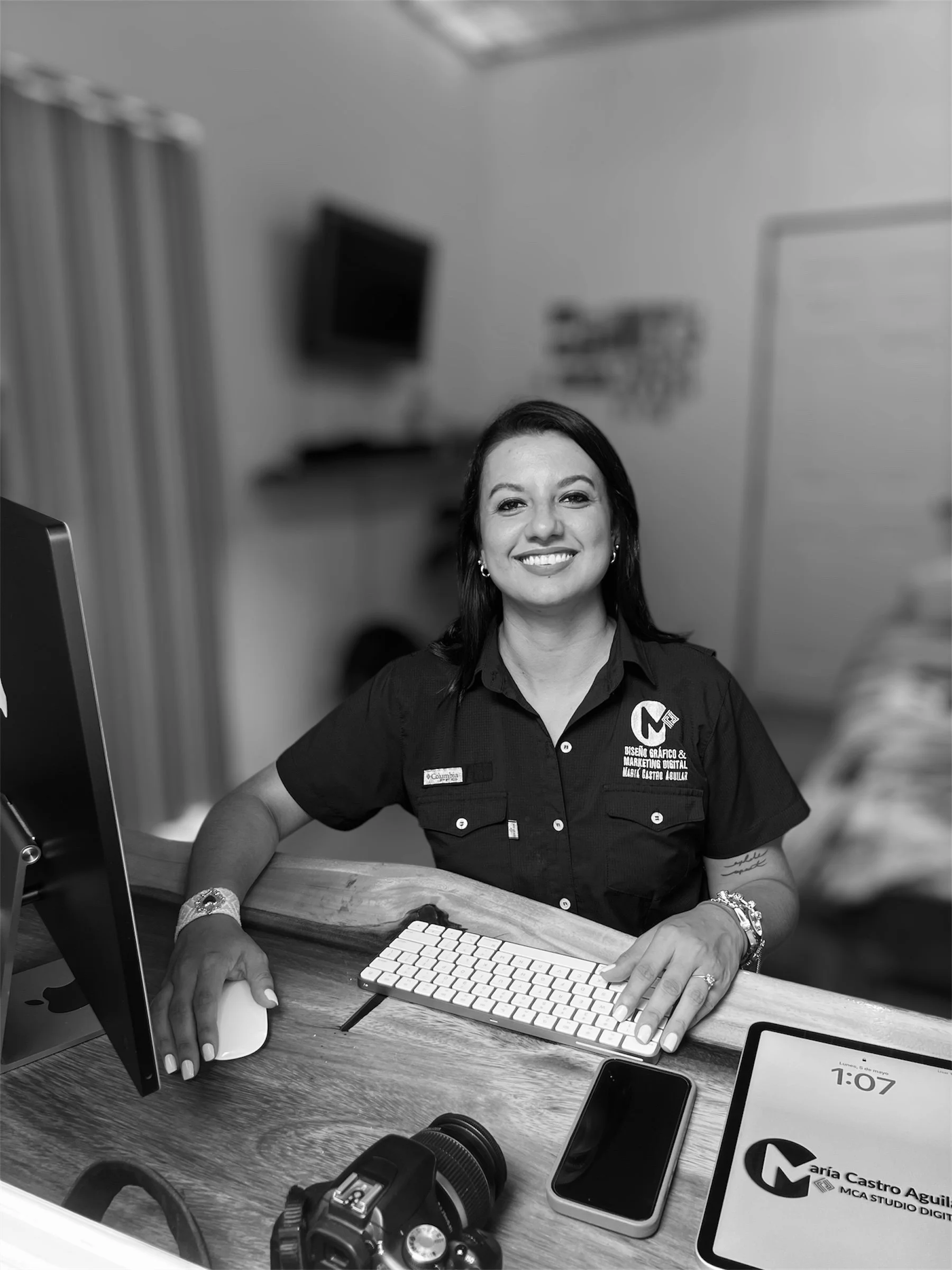Mujer sonriendo, sentada en una mesa de trabajo con computadora, teclado, teléfono móvil y cámara fotográfica, en un entorno de oficina