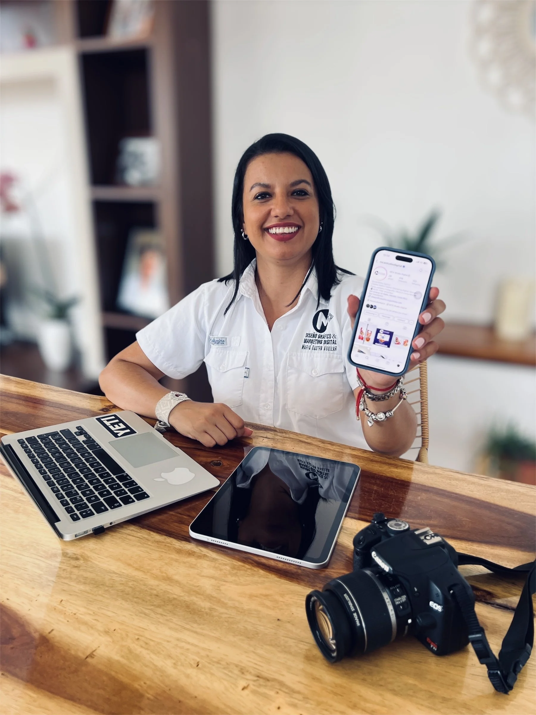Mujer sonriente mostrando un teléfono móvil con una pantalla visible en una mesa de madera con una laptop, una tableta y una cámara fotográfica.