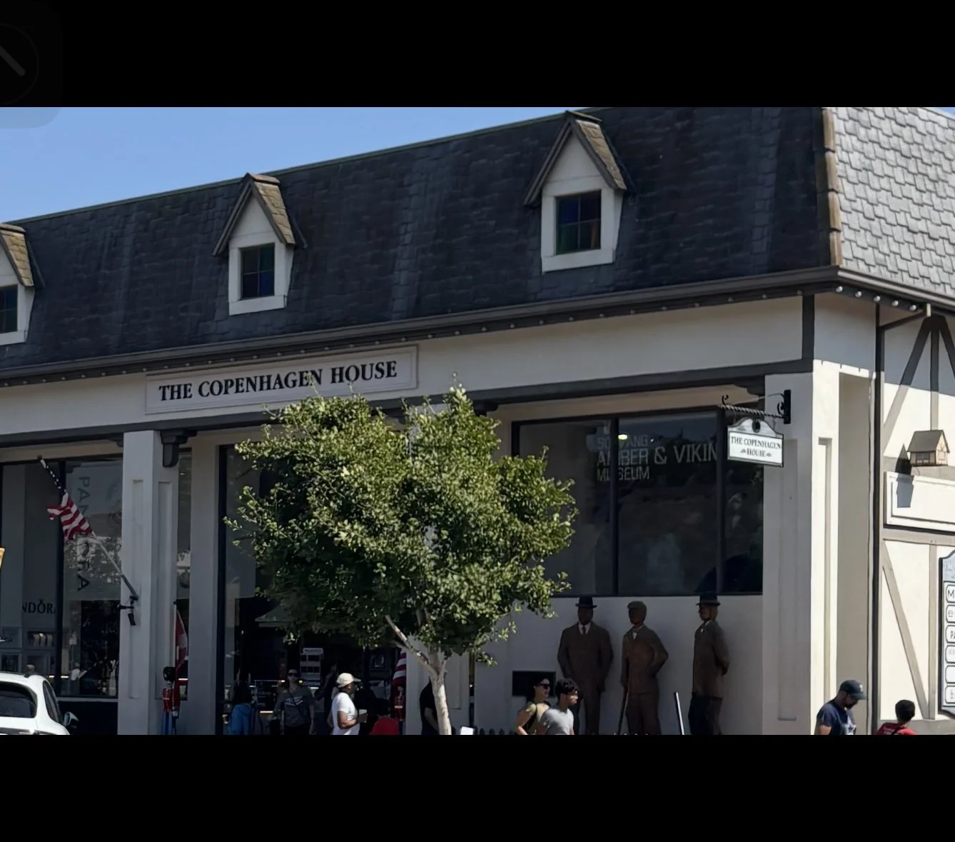 The Copenhagen House building with a black shingle roof, white walls, three small dormer windows, a sign reading 'The Copenhagen House', and statues of historical figures outside. Several people are walking in front.