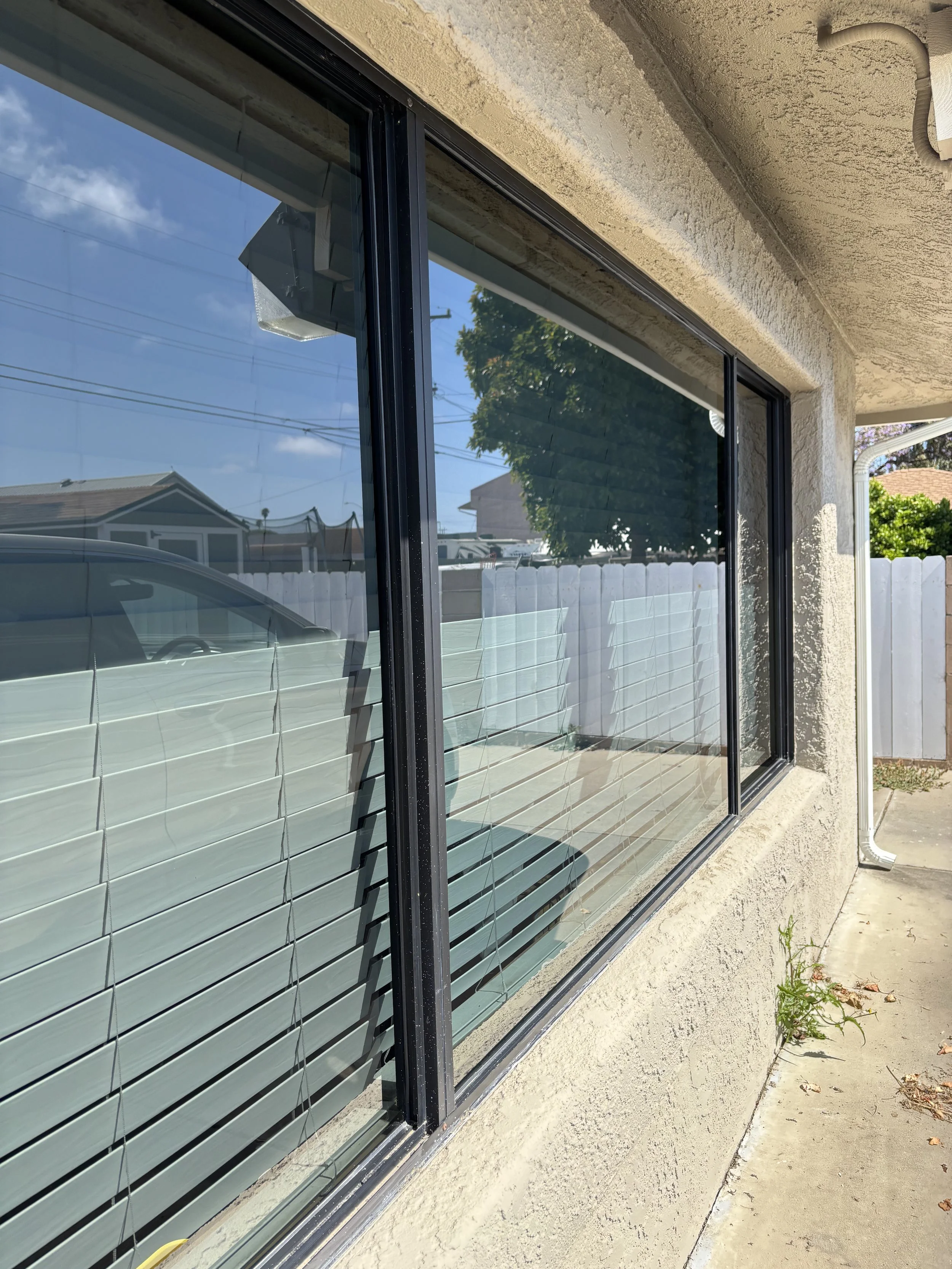 Exterior view of a building with a large window with blinds, beige stucco wall, and a concrete sidewalk with a small plant growing.