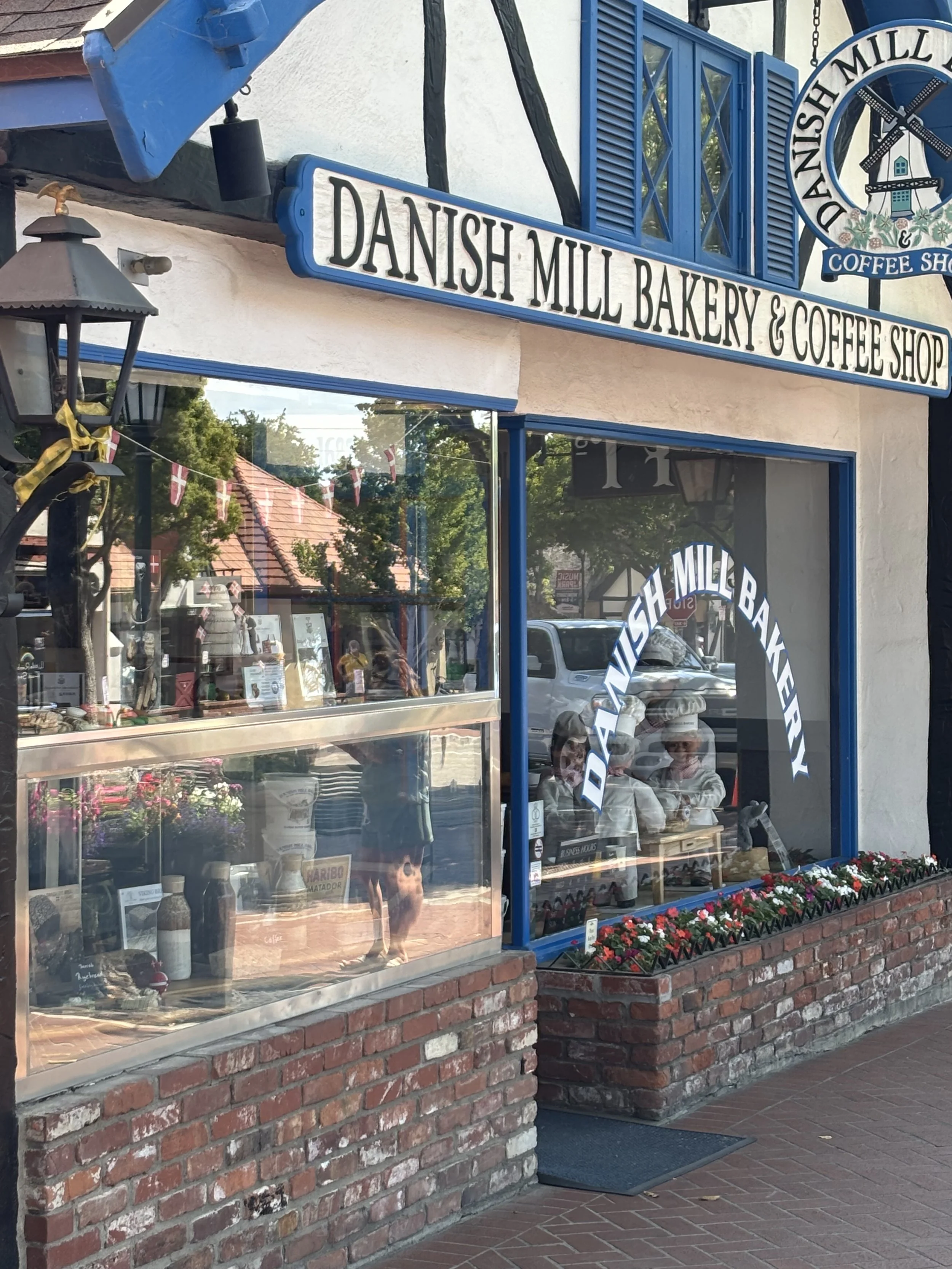 The exterior of Danish Mill Bakery & Coffee Shop with a signboard and brick window sills, decorated with flowers outside.
