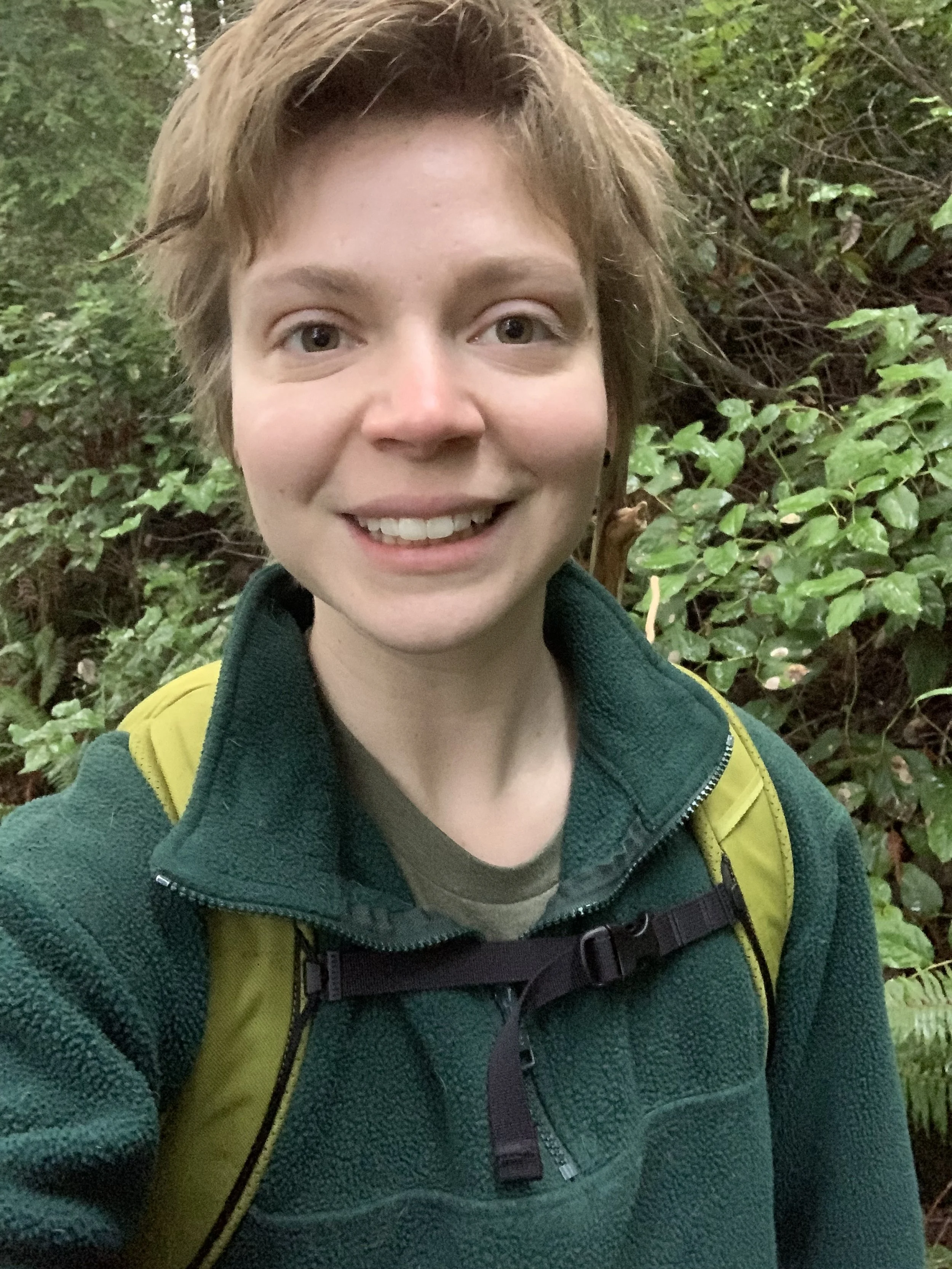 A person with blondish hair smiling outdoors in a green forest, wearing a green fleece jacket and a green backpack.