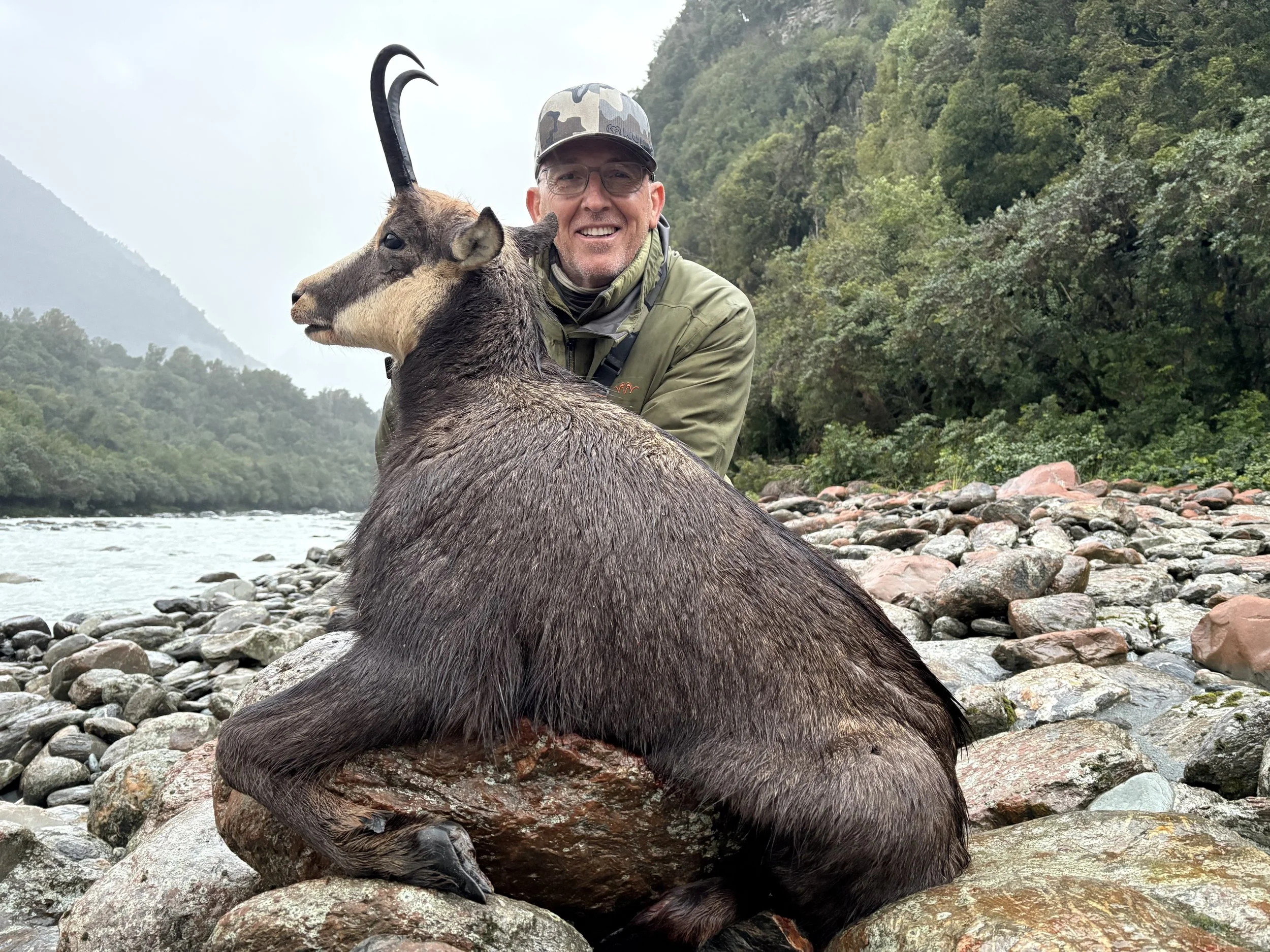 A man with glasses and a woodland camouflage cap smiling, sitting behind a large chamois , on a rocky riverbank with lush green trees and misty mountains in the background.