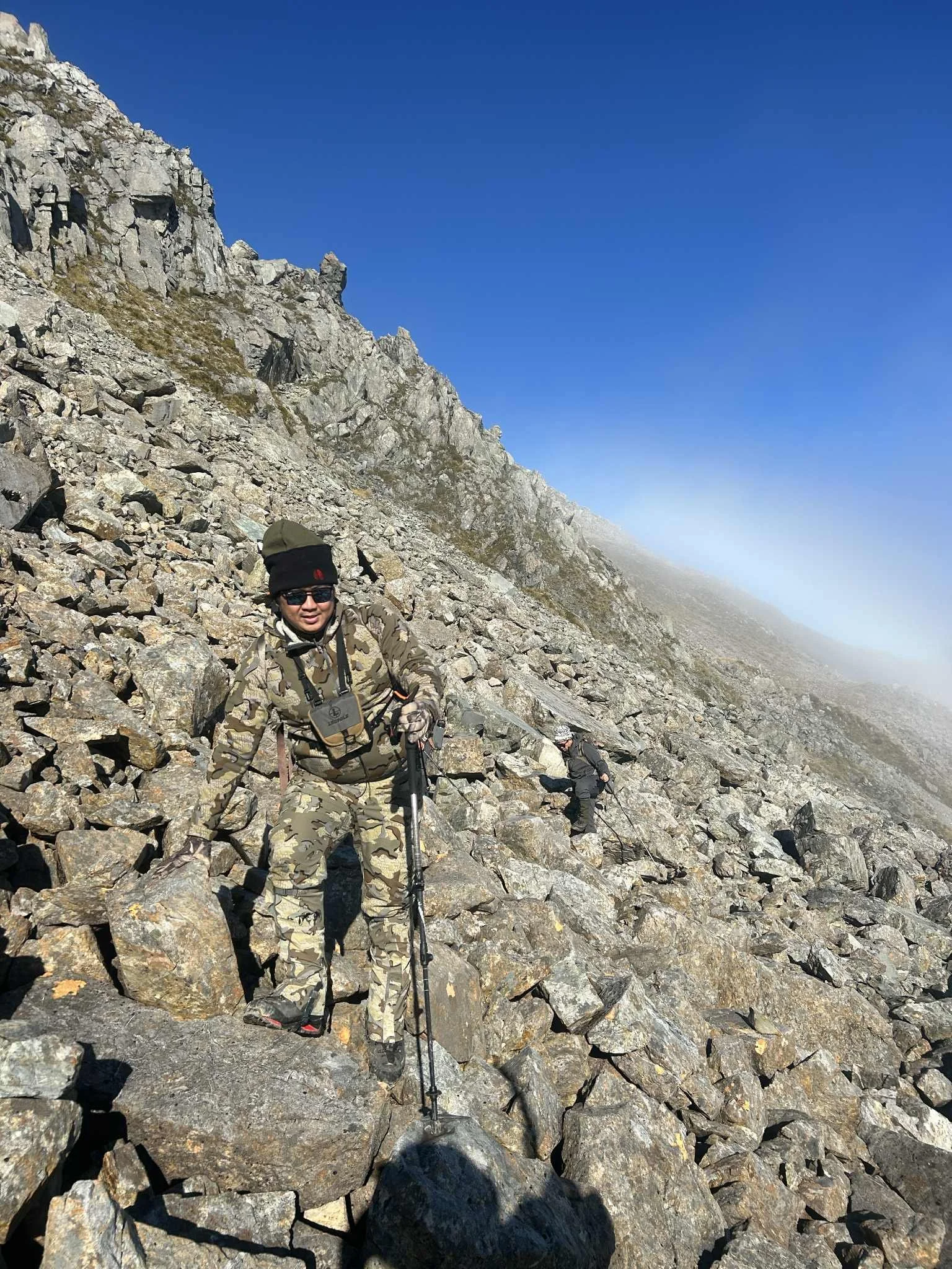 Two hikers in camouflage clothing climbing a rocky mountain slope under a bright blue sky, with some fog in the distance.
