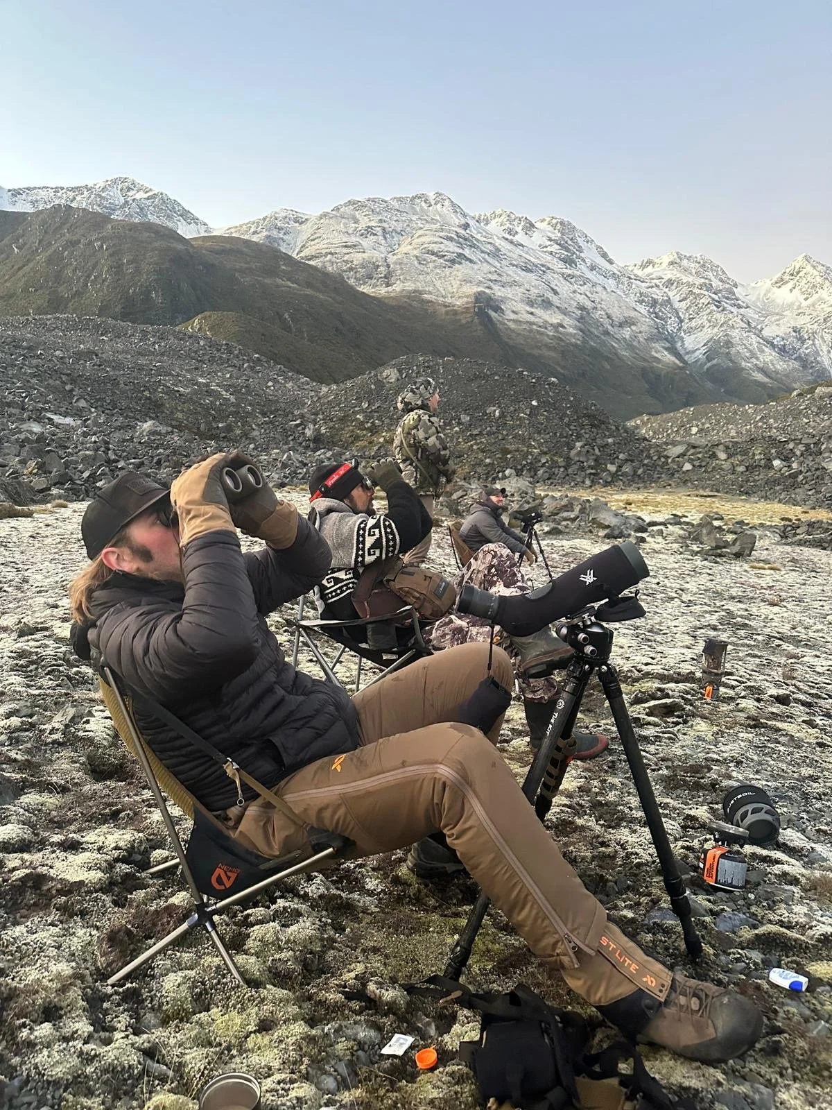 Group of people outdoors in a rocky, mountainous landscape, some looking through binoculars or binoculars, with a telescope set up on a tripod, during daytime.