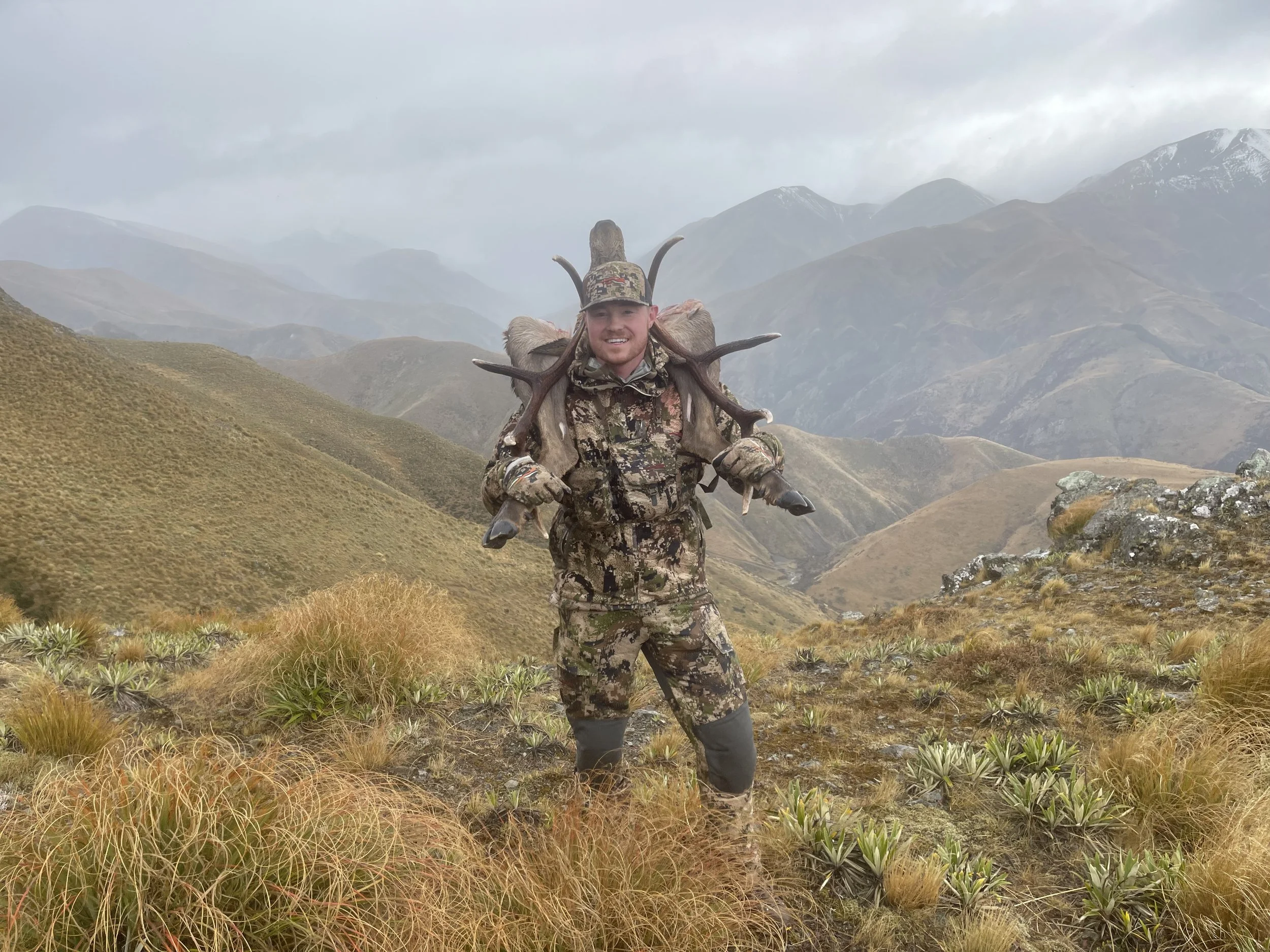 A man in camouflage hunting gear standing in a mountainous landscape, carrying animal antlers and fur on his back, smiling at the camera.