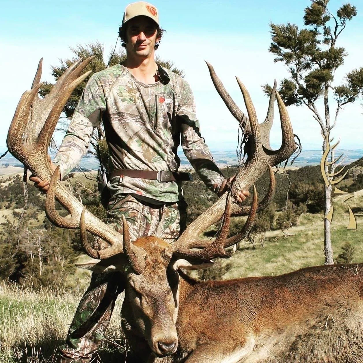 A man in camouflage clothing stands outdoors with a large red stag, holding its antlers in both hands. The deer is lying on the ground with a scenic landscape in the background.
