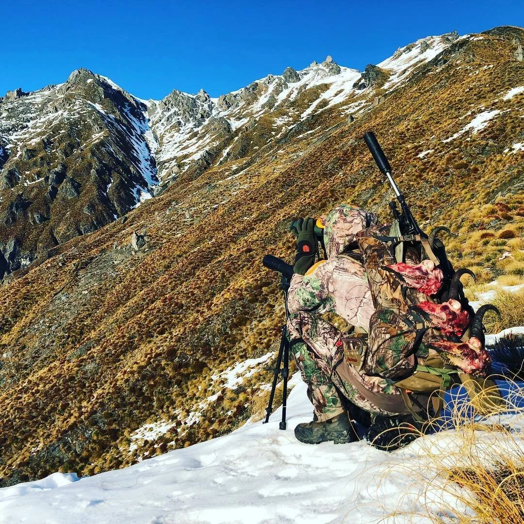 Person dressed in camouflage hunting gear, lying prone with a rifle on a mountain slope with snow patches, dry grass, and steep rocky peaks in the background.
