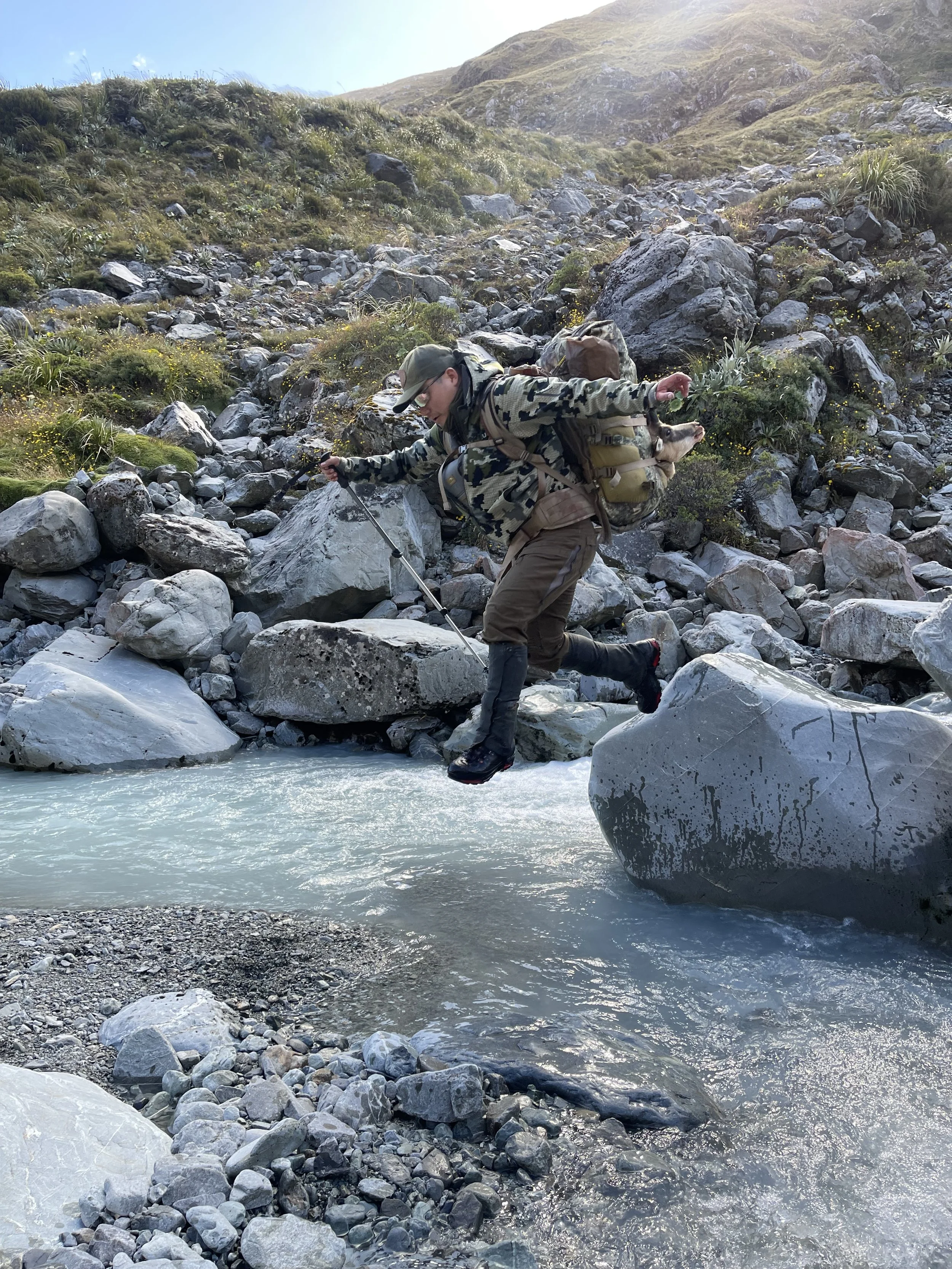 A person with a backpack jumping over a rocky stream in a mountainous landscape, accompanied by a small dog in the backpack.