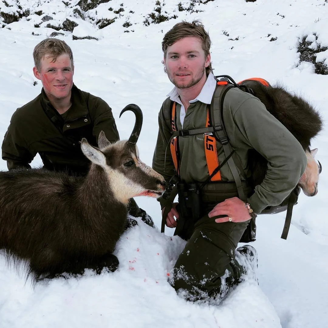 Two men in winter clothing kneeling in snow with a chamois buck, surrounded by snow-covered landscape.