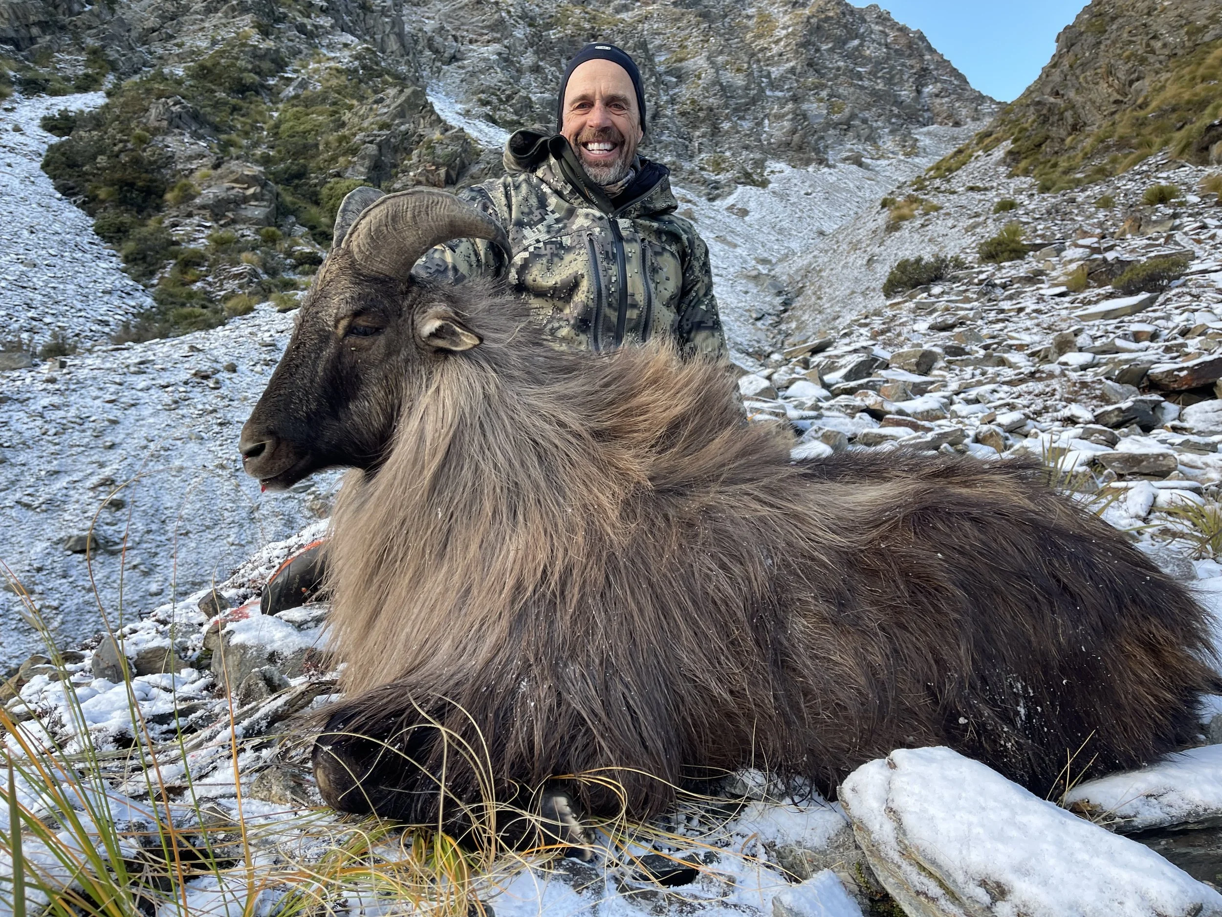 A man in camouflage gear smiling next to a large, fallen mountain goat with dark fur and curved horns in a snowy mountainous landscape.