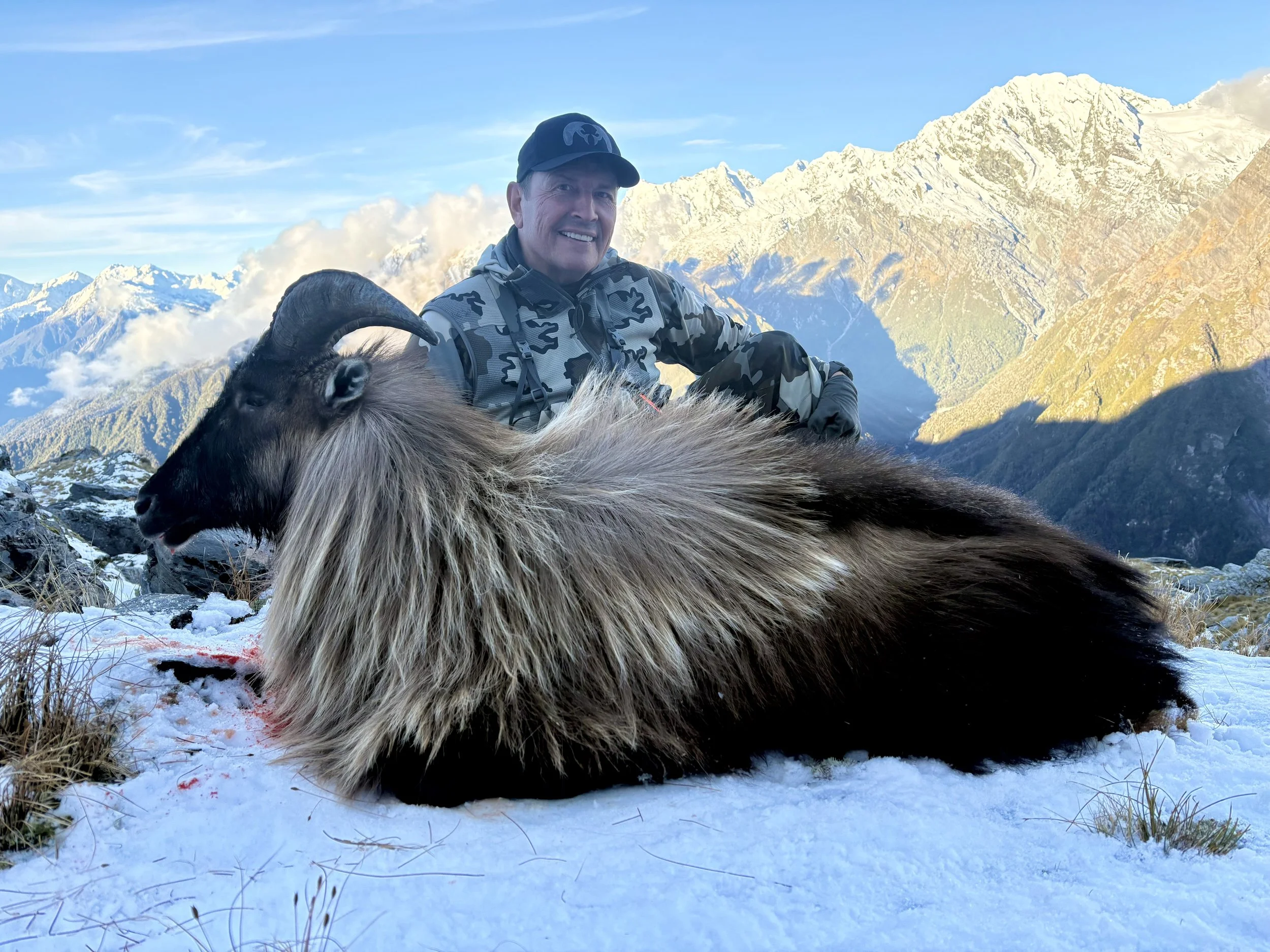 A man in outdoor camouflage gear smiling and sitting on snow-covered ground with a goat that has large curved horns, in a mountainous area with snow-capped peaks and a clear blue sky in the background.