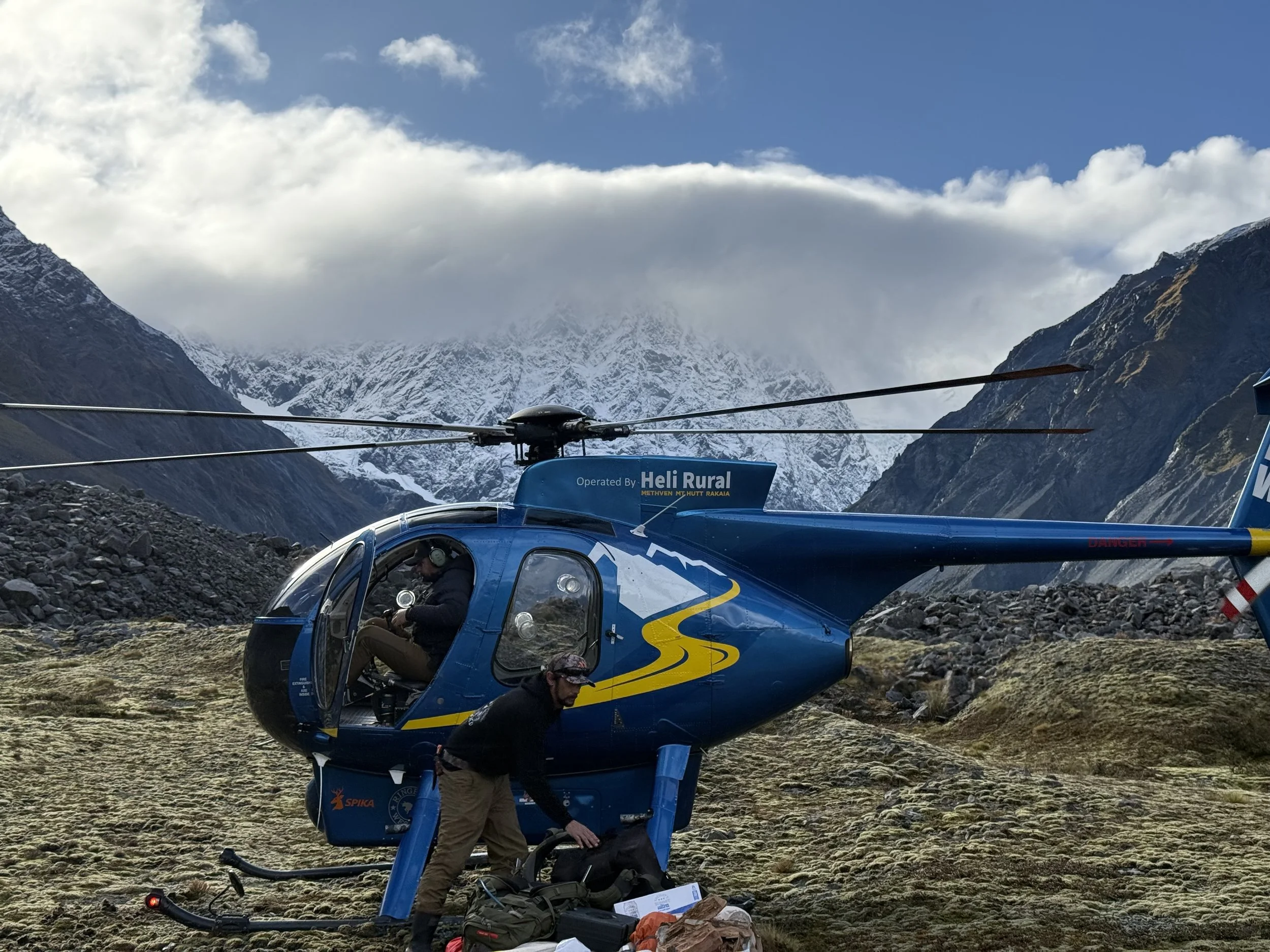 A blue helicopter on a grassy terrain in a mountainous landscape with snow-capped peaks and a cloudy sky in the background.