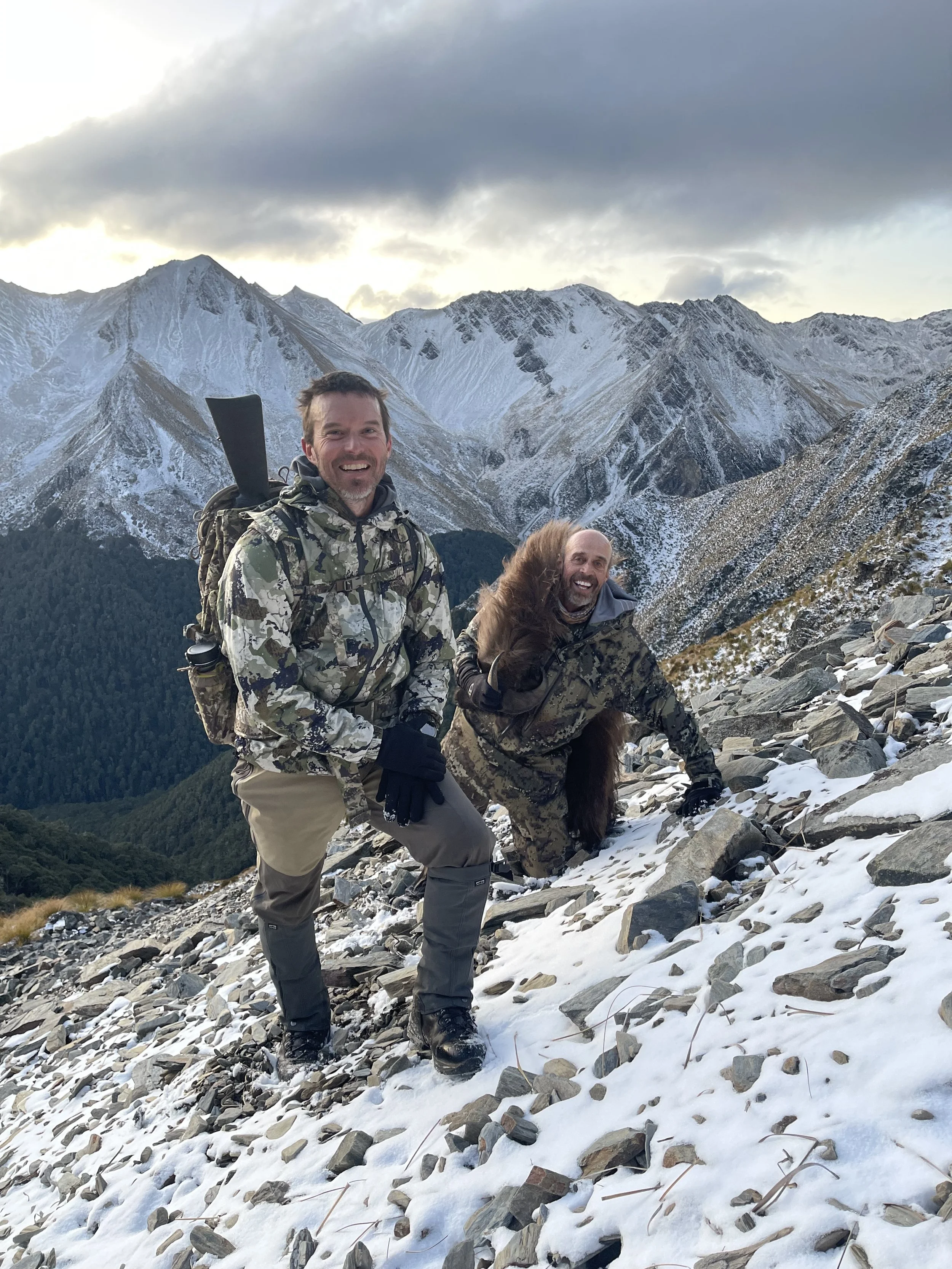 Two men in camouflage clothing hiking on a snowy mountain slope with a backdrop of snow-capped peaks and cloudy sky, smiling and posing for the photo.