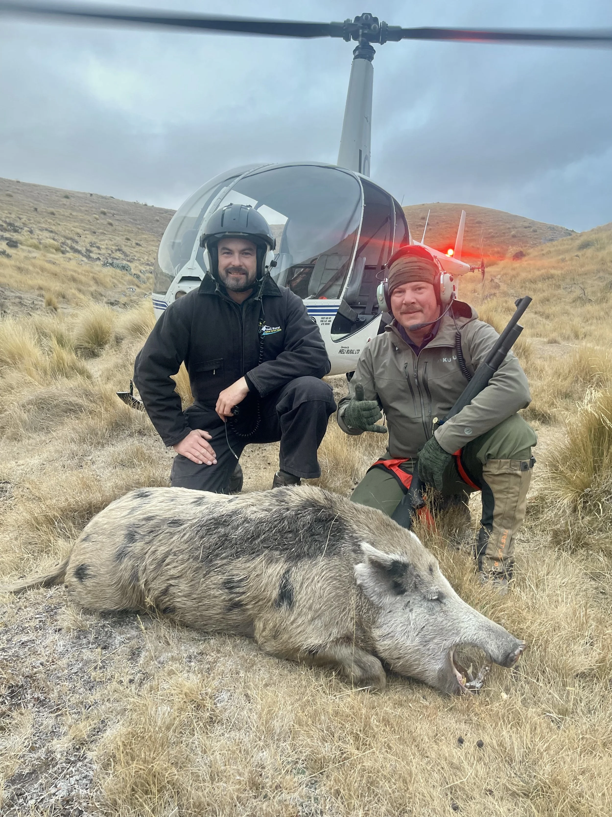 Two hunters in outdoor gear kneeling next to a dead wild boar on a grassy hillside, with a helicopter in the background.