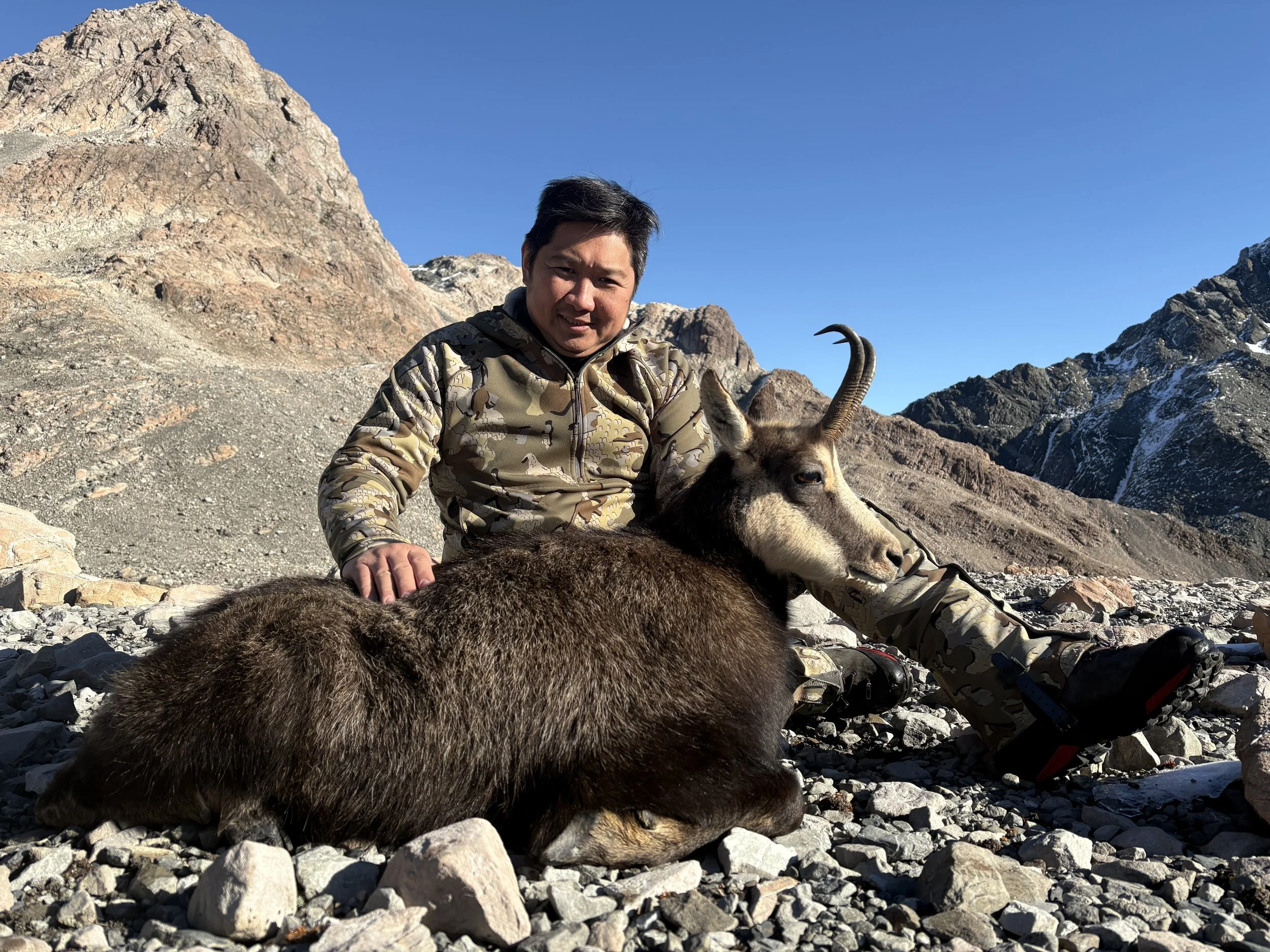 A man in camouflage clothing sitting on rocky terrain next to a goat, with mountains and a clear blue sky in the background.