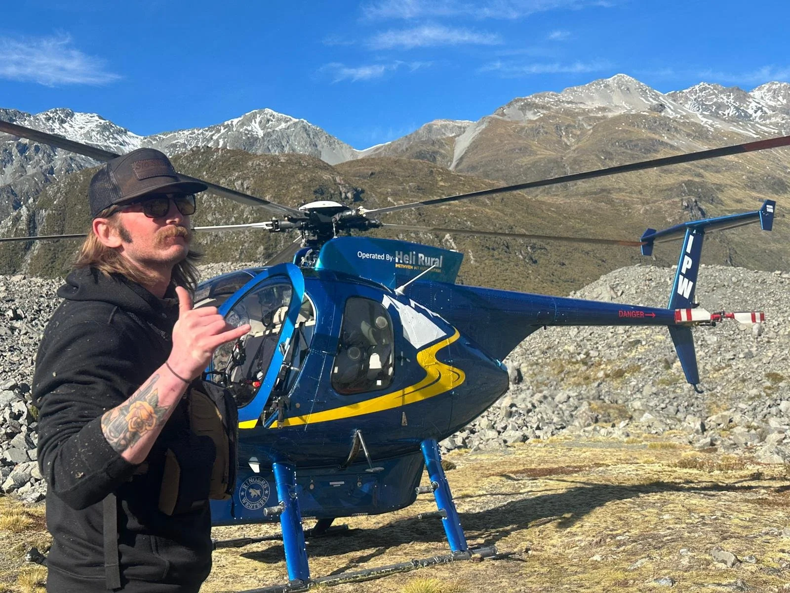 A man with long hair, sunglasses, and tattoos on his arm making a shaka sign next to a blue helicopter with mountain scenery in the background.